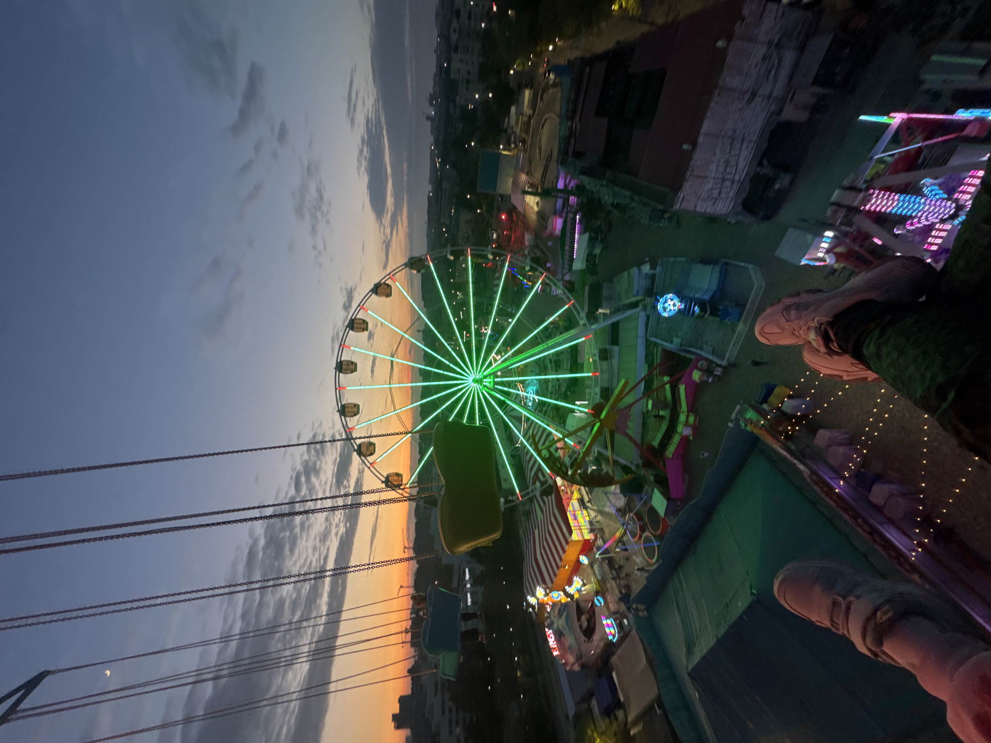 The image shows a tilted view from a high vantage point, likely a carnival ride, overlooking an amusement park in Yevpatoriya, Ukraine.
A Ferris wheel illuminated with green lights is a central focus. The sky is a blend of orange and blue at dusk with some clouds. A swing carousel with seats on chains is also visible. Carnival attractions and buildings, including an elevated green structure, fill the background.
In the foreground, the edge of the swing carousel and a person's legs and shoes are visible, suggesting the perspective is from someone on the ride.