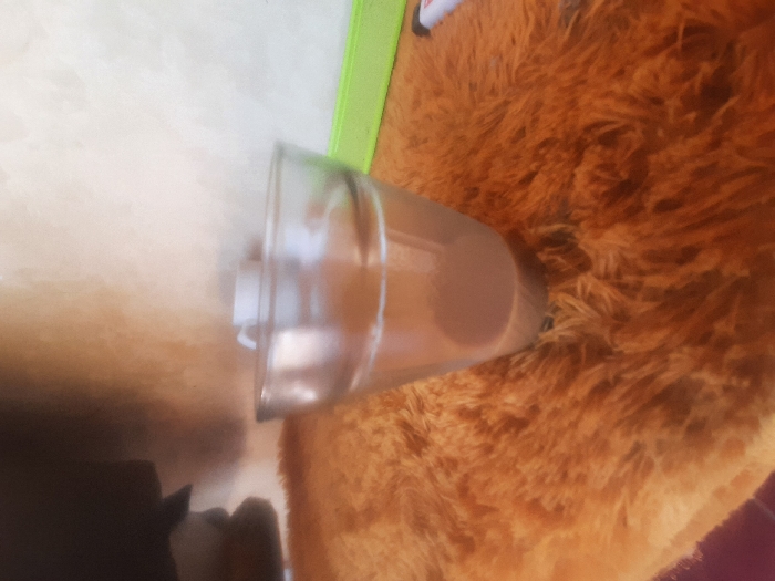 The scene depicts an indoor environment in Bandar Lampung, Indonesia. A transparent glass, approximately two-thirds full of a light brown liquid, likely coffee or tea, is centrally positioned on a textured, shaggy orange-brown surface, resembling a rug or blanket. In the background, an off-white and light brown wall is visible. To the upper right of the glass, a white ceramic cup is partially observed. Further to the right, a bright green horizontal strip or object is present, and a small white object with a red tip, possibly a marker or lighter, is partially visible at the far right. No individuals are present in the frame, and no specific actions or interactions are depicted.
