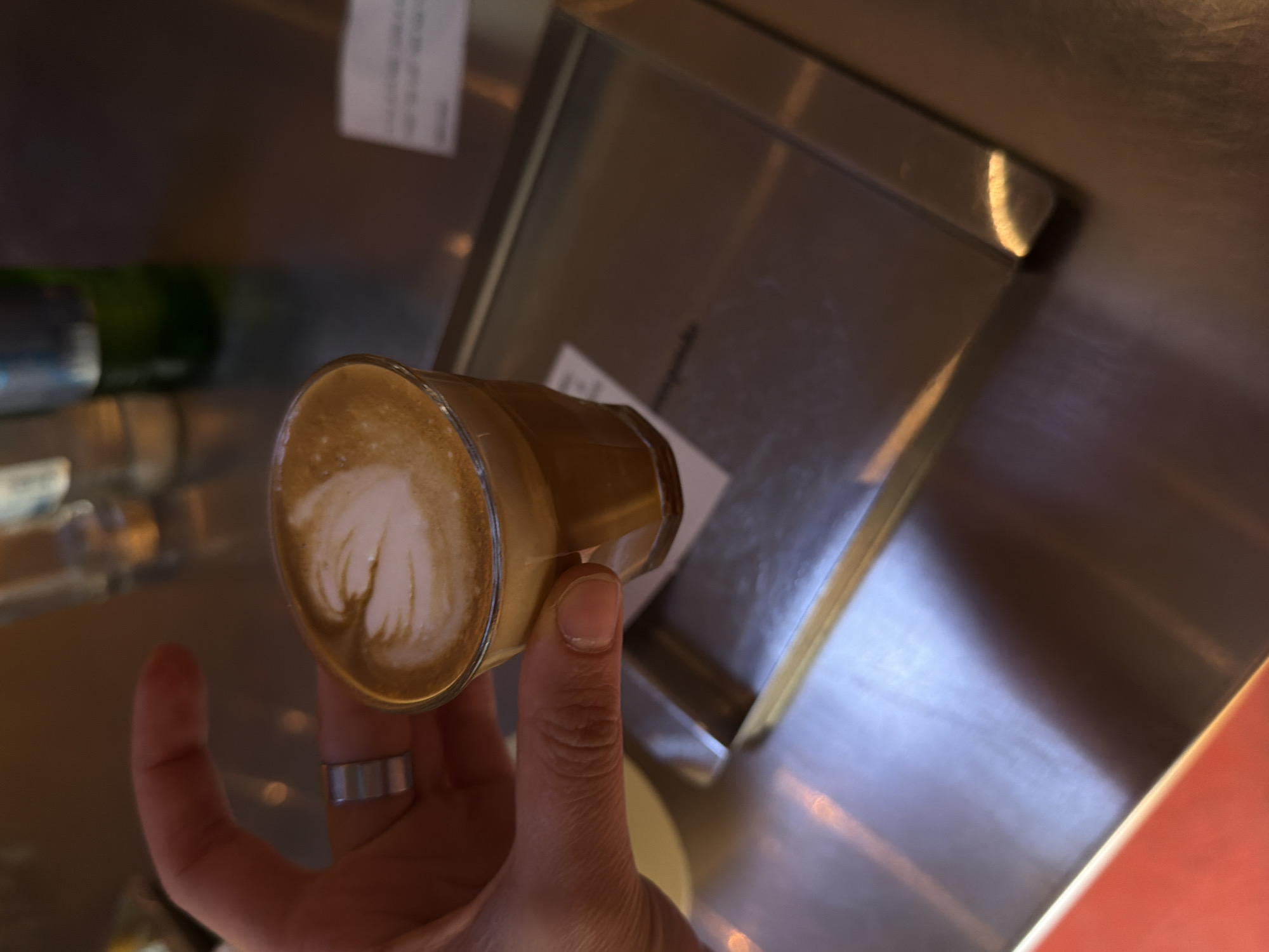 A hand holding a glass of coffee with latte art is depicted in the image. The hand has a ring on one of the fingers. The coffee glass appears to be a small cup or demitasse, and it's filled with a latte or cappuccino, the surface of which shows a milky design. The background is a neutral, metallic surface.
It's not possible to identify the exact location in Yerevan, Armenia, based solely on this image. More contextual details or landmarks would be needed to determine the location.