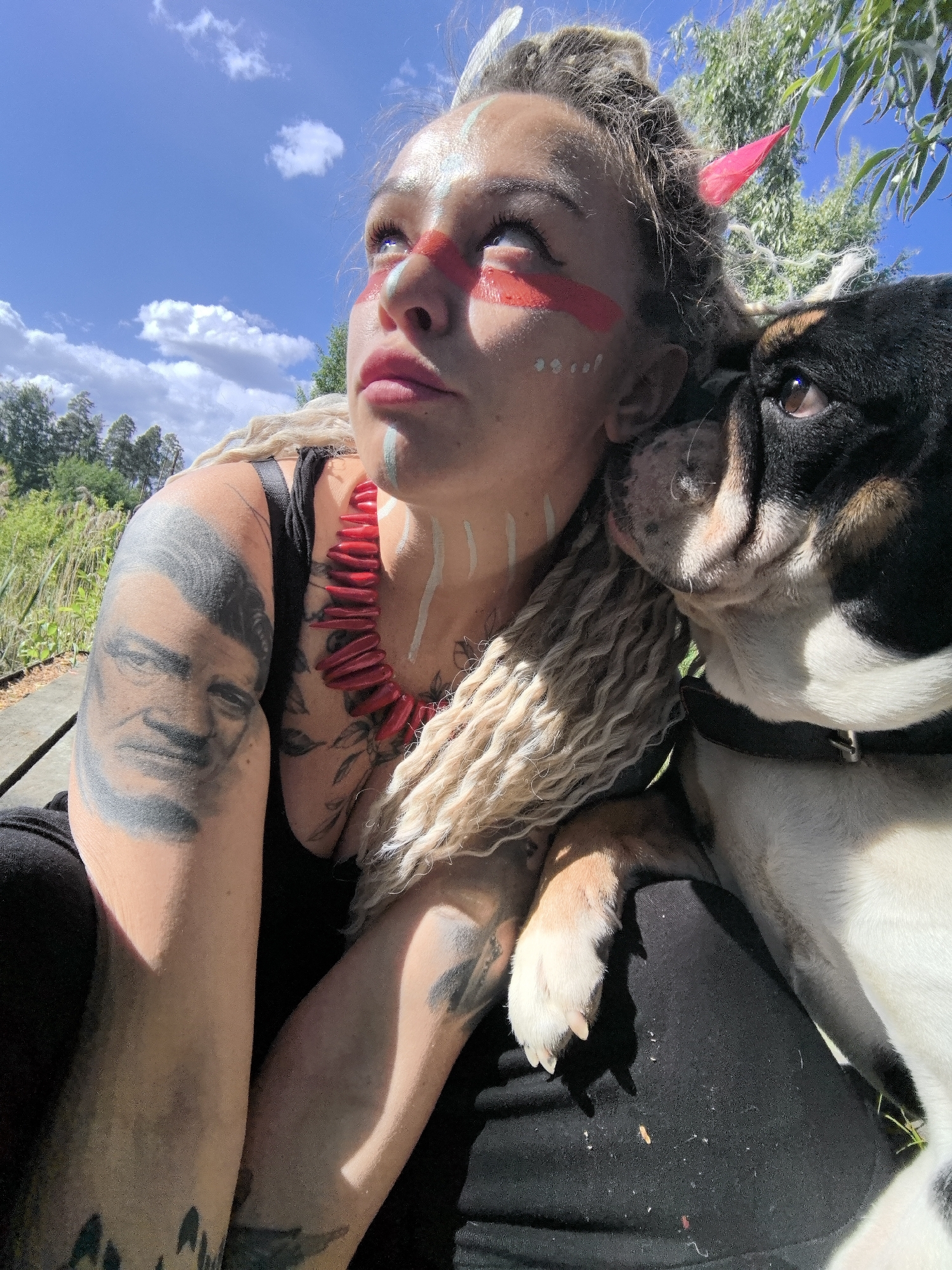 A young woman with face paint is outdoors with her bulldog on a sunny day. The woman has red and white face paint in stripes and dots. She has blonde dreadlocks and a red beaded necklace. She wears a black tank top, and has tattoos on both arms, one appearing to be a portrait. The bulldog, white with black markings, nestles its head against her cheek and rests its paw on her leg. The background reveals a partly cloudy sky, green trees, and possibly a garden area. The location of Noviy, Russia is not identifiable in the image.