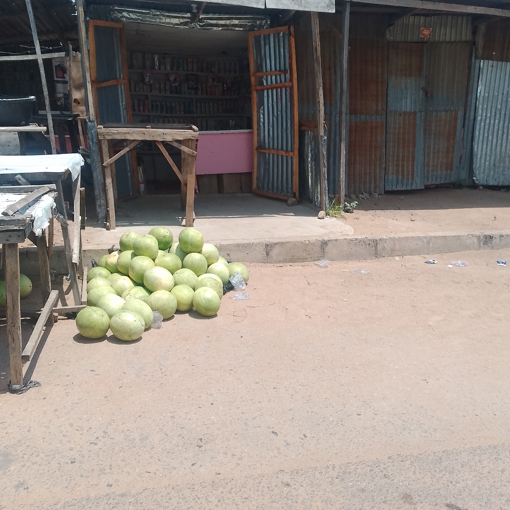 The image depicts an outdoor scene in Jalingo, Nigeria, likely a market or roadside stall. In the foreground, a large pile of green watermelons is displayed on the sandy ground. To the left, a makeshift wooden structure, possibly a table or stall, partially obscures the view. Behind the watermelons and the structure is a shop with corrugated metal walls and a wooden-framed door, partially open, revealing shelves stocked with various items. The ground appears to be a dirt or paved road with some scattered plastic debris. The overall impression is one of a typical street vendor setup in a developing urban environment.