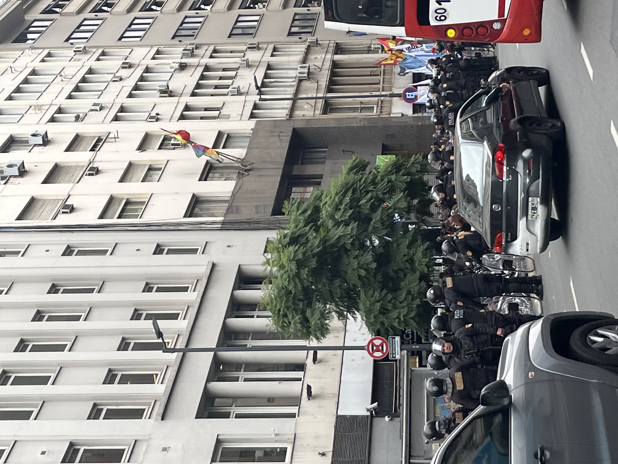 The image shows a street scene in Buenos Aires, Argentina. There are several tall buildings on the left side of the frame. A line of police officers in riot gear is standing on the right side of the street, next to several cars and a red bus. There is a tree in the middle of the frame. A colorful kite is flying near the top of the buildings.