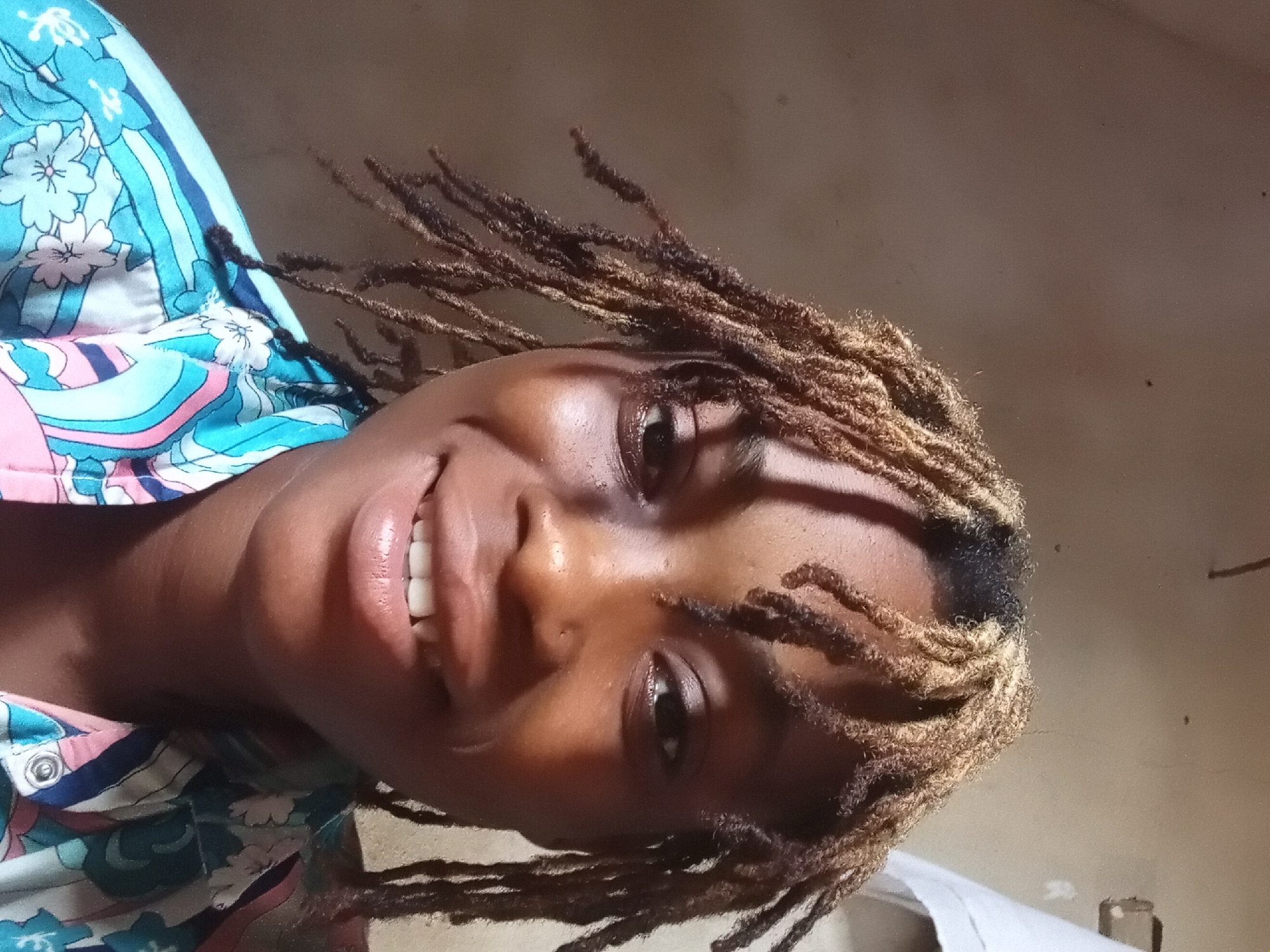 The image shows a smiling person with dreadlocks, wearing a patterned shirt with blue, pink, and white colors. The background is indistinct but appears to be a simple interior wall. The location is Unknown, Unknown.