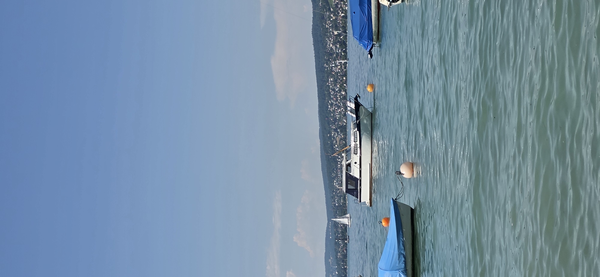This is a vertical, eye-level shot of Lake Zurich, near Oberrieden, Switzerland, on a clear, sunny day. The water is a calm, turquoise color with small ripples. In the foreground, two boats are moored. The boat on the left is covered with a blue tarp. In the middle ground, a white motorboat with a blue canopy is anchored, and a yellow buoy floats nearby. Further out on the lake, a white sailboat can be seen. The background features a densely populated shoreline with buildings and lush green trees, characteristic of the Oberrieden area. The sky is a clear, light blue with a few wispy clouds. There are no people visible in the image.