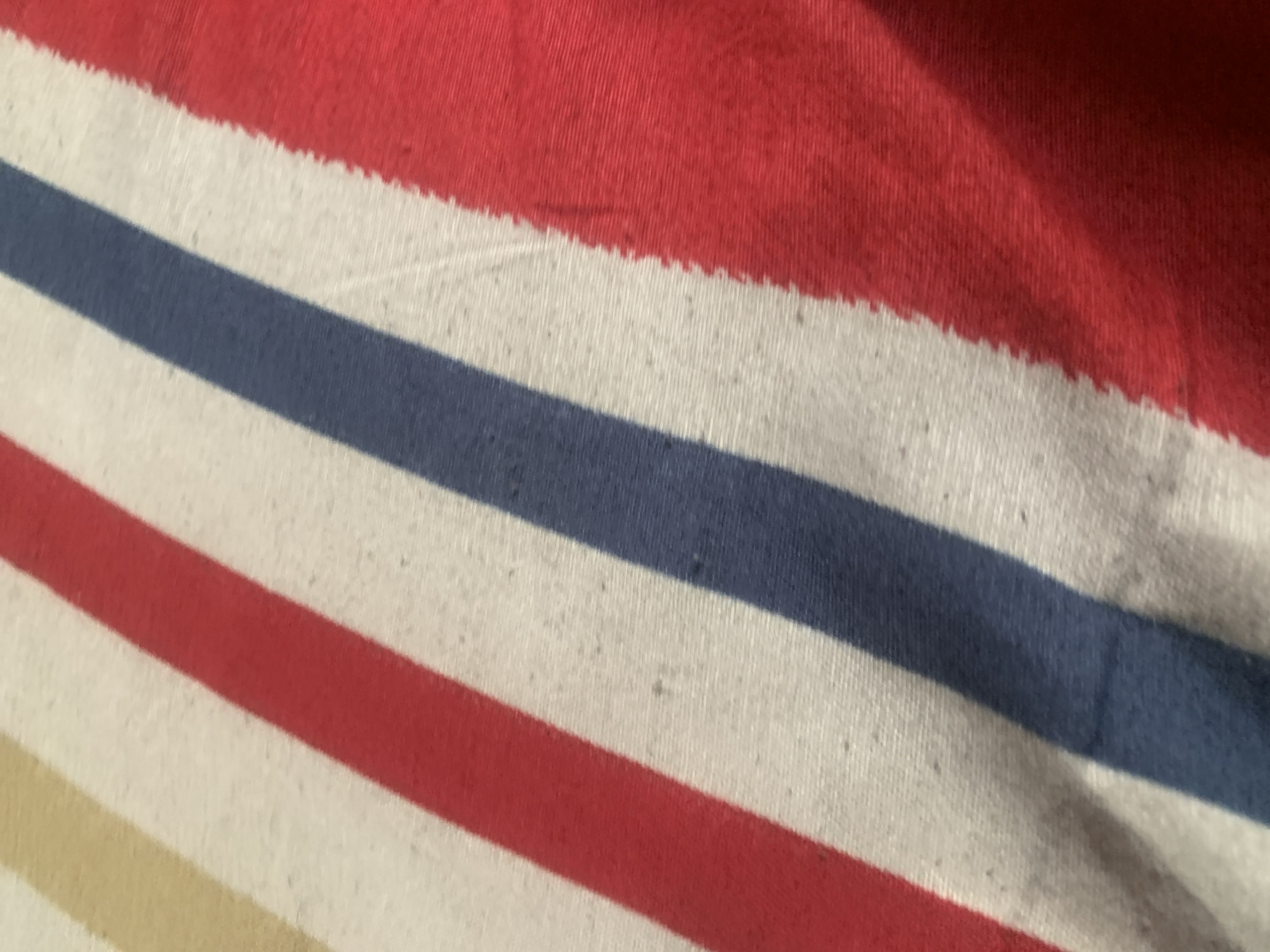 The image shows a close-up, diagonal view of a striped fabric. The stripes are red, white, and blue, with a hint of a tan or yellow stripe at the top left. The fabric appears to be a woven material, possibly a blanket or a tablecloth, with visible texture and some minor imperfections such as small dark specks. There are no people, identifiable landmarks, or contextual environmental details present in the image. Therefore, it is impossible to determine the location of this image within the city of Jauro Yinu, Nigeria.