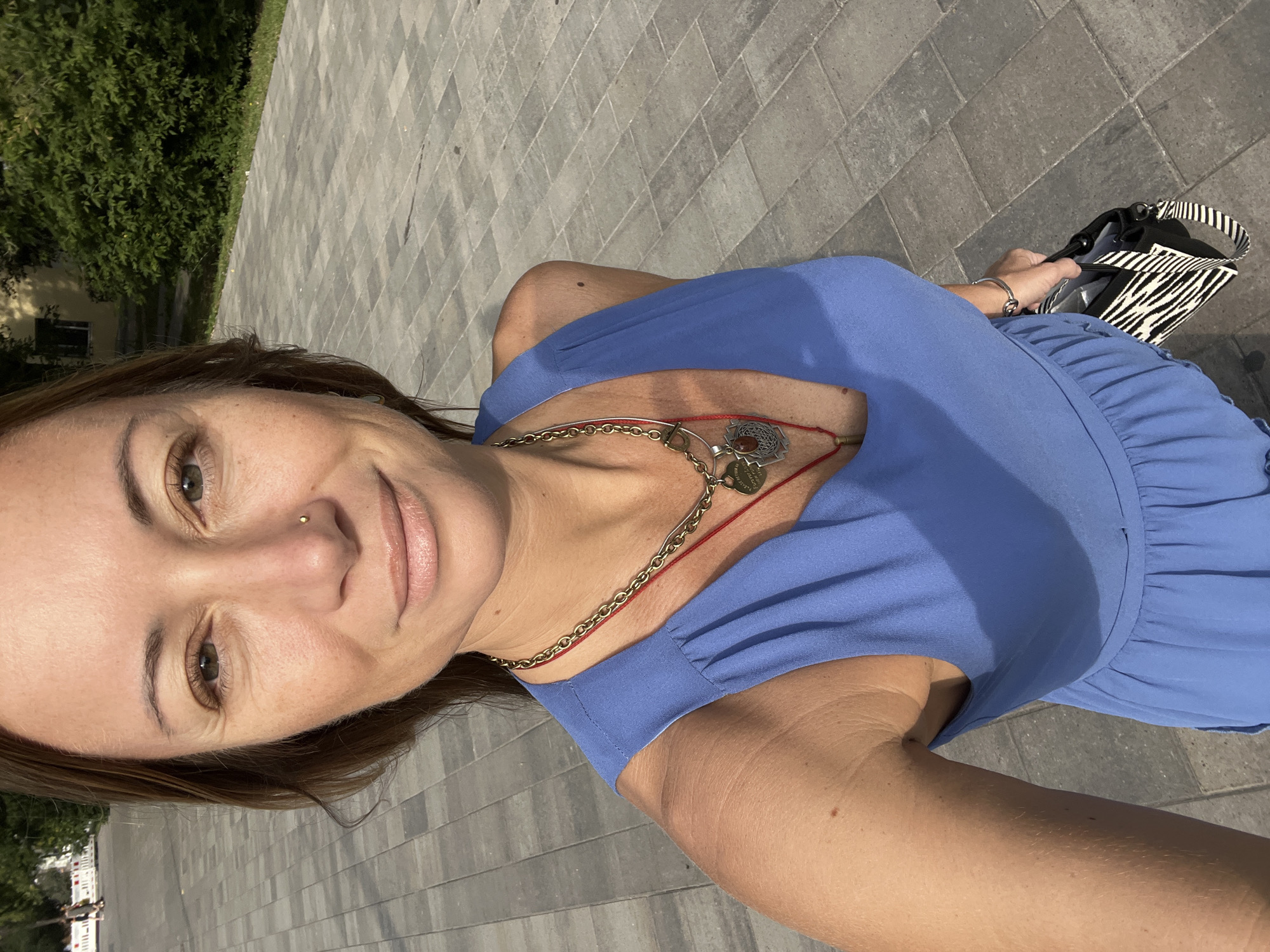 A medium shot shows a woman from the chest up, taking a selfie outdoors. She has long brown hair and is wearing a blue dress and a necklace. The background shows a paved walkway and some trees. The scene is set in the city of Novokuybyshevsk, Russia.