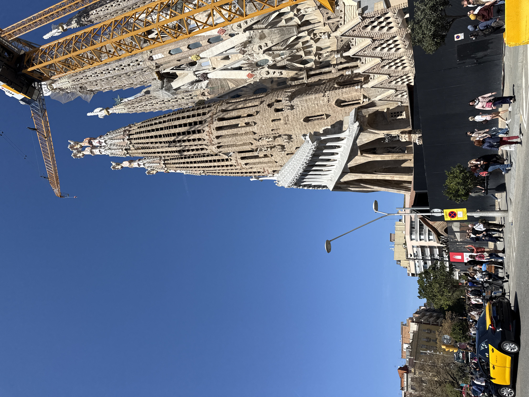 The image shows the Sagrada Familia basilica in Barcelona, Spain. The basilica's facade is partially visible, along with a large yellow crane involved in its ongoing construction. The sky is clear and blue. A crowd of people is present on the sidewalk adjacent to the building, and several pedestrians are walking by. There are trees and buildings on the opposite side of the street. A yellow and black taxi cab is parked along the sidewalk. A streetlight is visible.