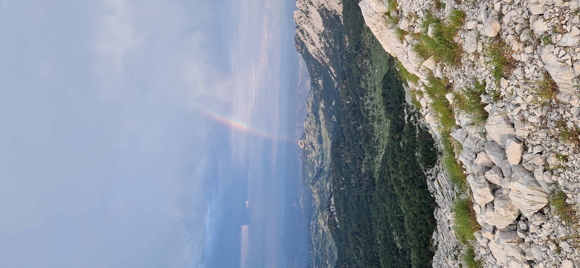 An outdoor, high-angle, vertical, full shot captures a scenic mountainous landscape under a cloudy sky, with a faint rainbow visible. The foreground is dominated by a rocky, uneven terrain covered in scattered grey rocks and patches of green grass. Beyond the immediate foreground, the terrain slopes down into a valley filled with dense, dark green forest. Further in the distance, rolling hills and mountains are visible, characterized by lighter rocky outcrops and vegetation. The sky is a pale blue, with scattered grey clouds, some of which appear heavy and dark, suggesting recent or impending rain. The rainbow, a subtle arc of colors, appears in the middle distance, emerging from behind a dark cloud and arching across the sky. The overall scene depicts a natural, rugged environment, possibly in or near Ubla, Bosnia and Herzegovina, at a time when the weather is transitional. There are no people or discernible structures in the image.