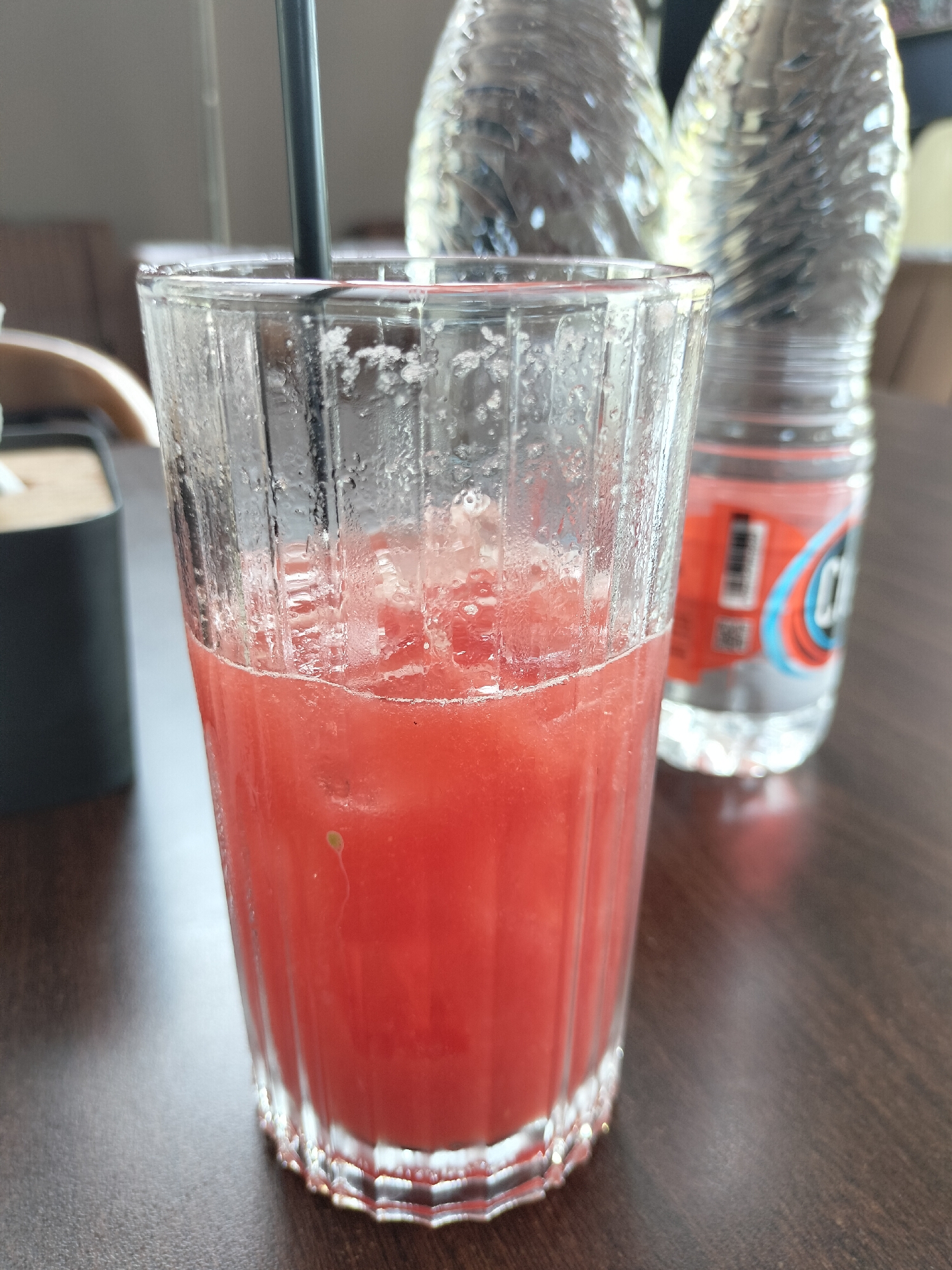 A glass of pink liquid, possibly juice, with ice and a black straw, sits on a dark brown wooden table. In the background, two plastic bottles of water are visible, one with a red and blue label. To the left of the glass is a black rectangular object, possibly a tissue holder. The scene appears to be indoors, perhaps at a cafe or restaurant, with a neutral-colored wall and furniture visible in the blurred background. The lighting suggests it is daytime. The image does not contain any information or visual cues that would place it specifically in the city of Idi Rayeuk, Indonesia.