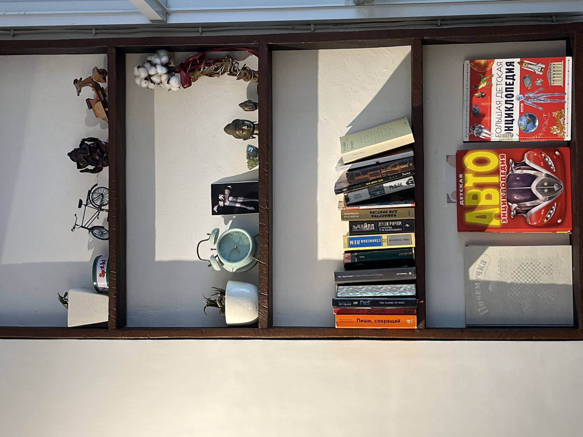 The image shows a wooden shelving unit against a white wall, likely indoors in Kuta Selatan, Indonesia. The unit contains various items arranged in three distinct sections.
On the left, there are wooden figurines, including an animal shape and a human figure, along with a bicycle model. A small can of "Spoti" is also visible.
The middle section features decorative elements like cotton bolls, possibly dried flowers or branches, and small Buddha-like statuettes. A black and white photo and a mint-green alarm clock are also placed there. A small white pot holds a plant.
The right section consists of stacked books, including Russian encyclopedias for children, with covers showcasing illustrations of vehicles and space exploration. Also among the books are literary works like "Wings of Fire" and "Ronin," and books on Kamasutra and other topics. "Почемучка" book is included, as well.