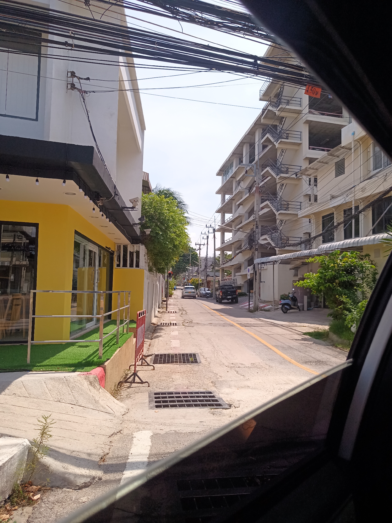 The image, taken from inside a vehicle, shows a street in Hua Hin City Municipality, Thailand. On the left is a yellow and white building with a black awning and artificial grass. A red safety barrier is placed along the sidewalk. The street has a yellow dividing line and several drainage grates. On the right is a tall, white building with exterior staircases. Several vehicles, including a black pickup truck and a scooter, are visible further down the street. Overhead, there are numerous tangled electrical wires. The sky is light blue.