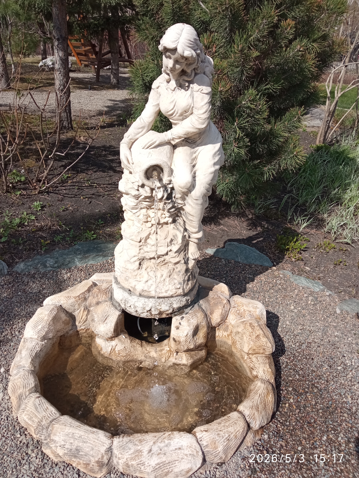 The image shows a white stone fountain with a statue of a woman pouring water from a jug into a stone basin. The fountain is surrounded by gravel. In the background, there are trees, including a coniferous tree, and some bare branches. The image was taken on May 3, 2026, at 3:17 PM. The location is Krasnoyarsk, Russia.