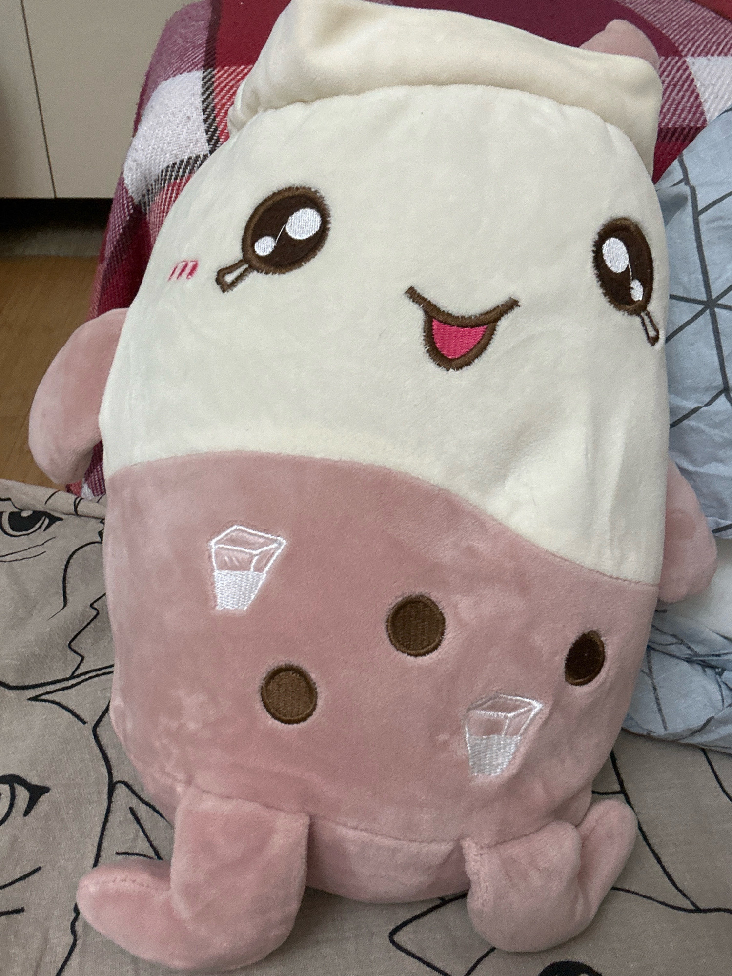 A plush toy, resembling a bubble tea drink, is lying on a bed. The toy is predominantly white and pink, with embroidered facial features including large brown eyes with white highlights, a pink mouth, and red blush marks on its cheeks. It has two brown circular "bubbles" embroidered on its lower pink section, and two small white embroidered cups with liquid are also visible on the pink section. The toy has small pink arms and feet. In the background, a red and white plaid blanket is visible, as well as a blue and gray patterned fabric. The setting appears to be indoors. No specific location in Khyrdalan, Azerbaijan, can be determined from the image.