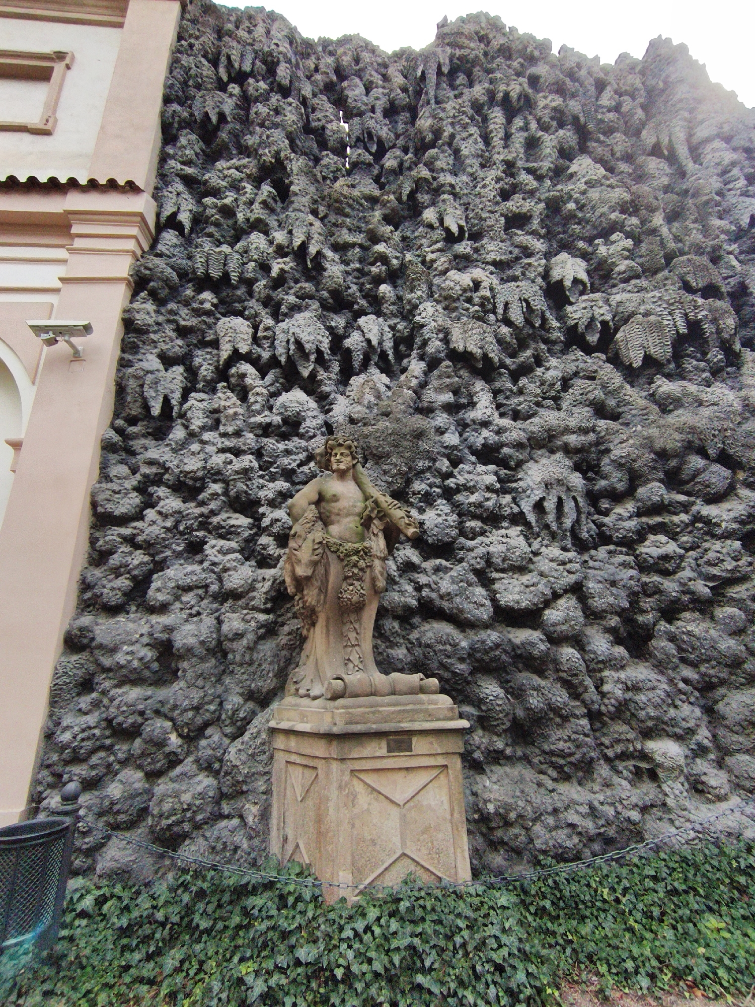 This is a photograph of the Wallenstein Garden, located in Prague, Czechia. The image depicts a striking grotto wall, a large, irregular, and textured rock formation that serves as a dramatic backdrop. In the foreground, a classical stone statue stands on a pedestal in front of the grotto. The statue appears to be a male figure, possibly a mythological character, adorned with drapery and foliage. The base of the statue is surrounded by lush green ivy, creating a vibrant contrast with the stone and rock. To the left of the scene, a portion of a light pink building with architectural details, including a window and decorative molding, is visible. A security camera is mounted on the building. A dark metal trash receptacle is positioned to the left of the statue. A metal chain forms a barrier in front of the ivy and the statue.
