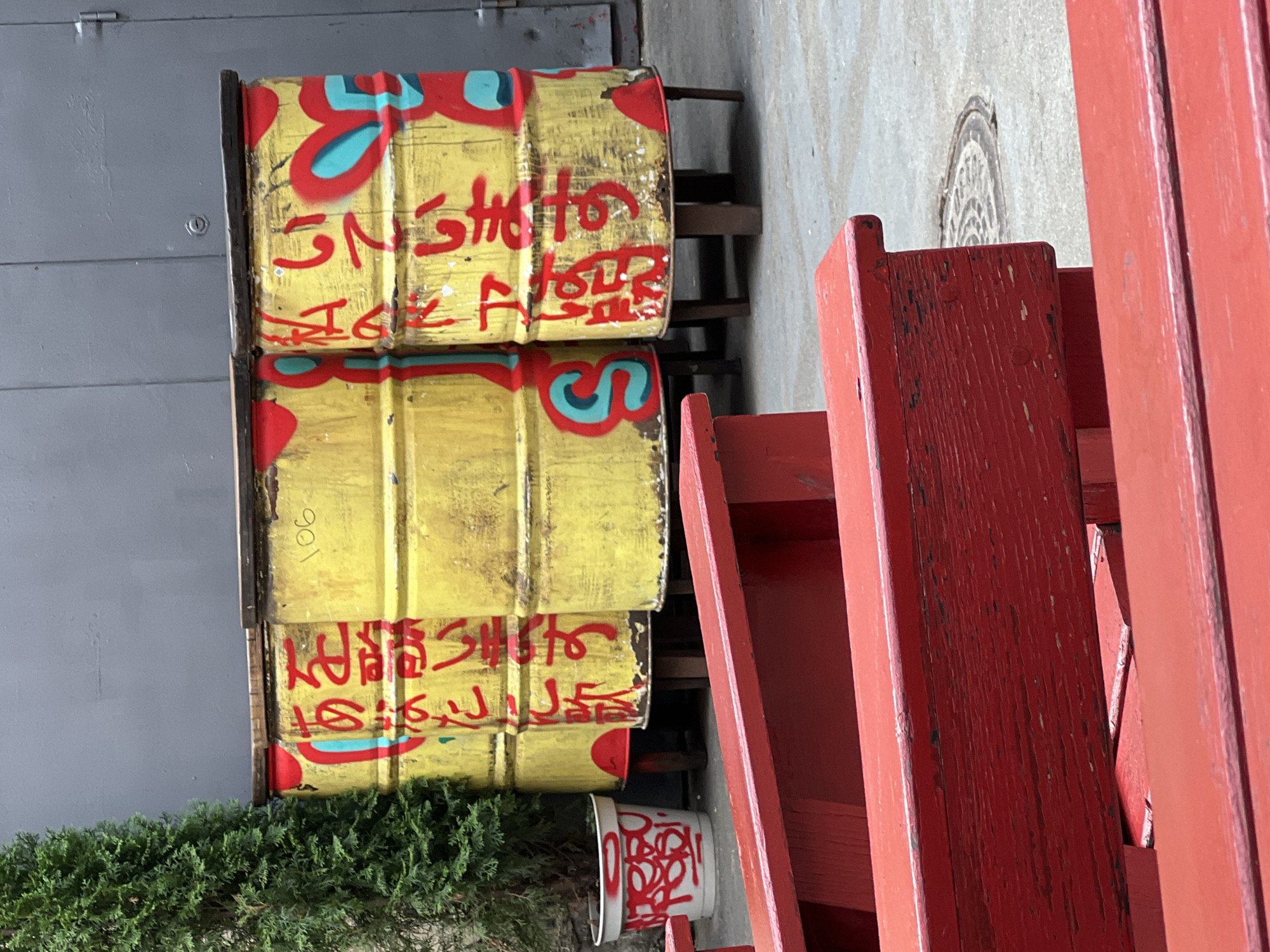 The image depicts an outdoor seating area with red wooden benches in the foreground, leading to three yellow oil drums with red and turquoise graffiti. The oil drums are standing on short wooden legs. The drum on the left has Japanese characters in red and a red and turquoise circular design. The middle drum has the number "106" written in yellow and red graffiti. The drum on the right also features Japanese characters in red and a red and turquoise heart-shaped design. To the left of the drums, a white planter with red graffiti is visible, containing a green coniferous plant. The background consists of a grey wall and a section of a metal door. The ground is paved with grey flagstones, and a manhole cover is visible in the lower right. The scene suggests an urban outdoor space, possibly a cafe or a public area in Moscow, Russia, given the presence of Japanese graffiti which might indicate a themed establishment or artistic expression common in certain city districts. There are no people visible in the image.