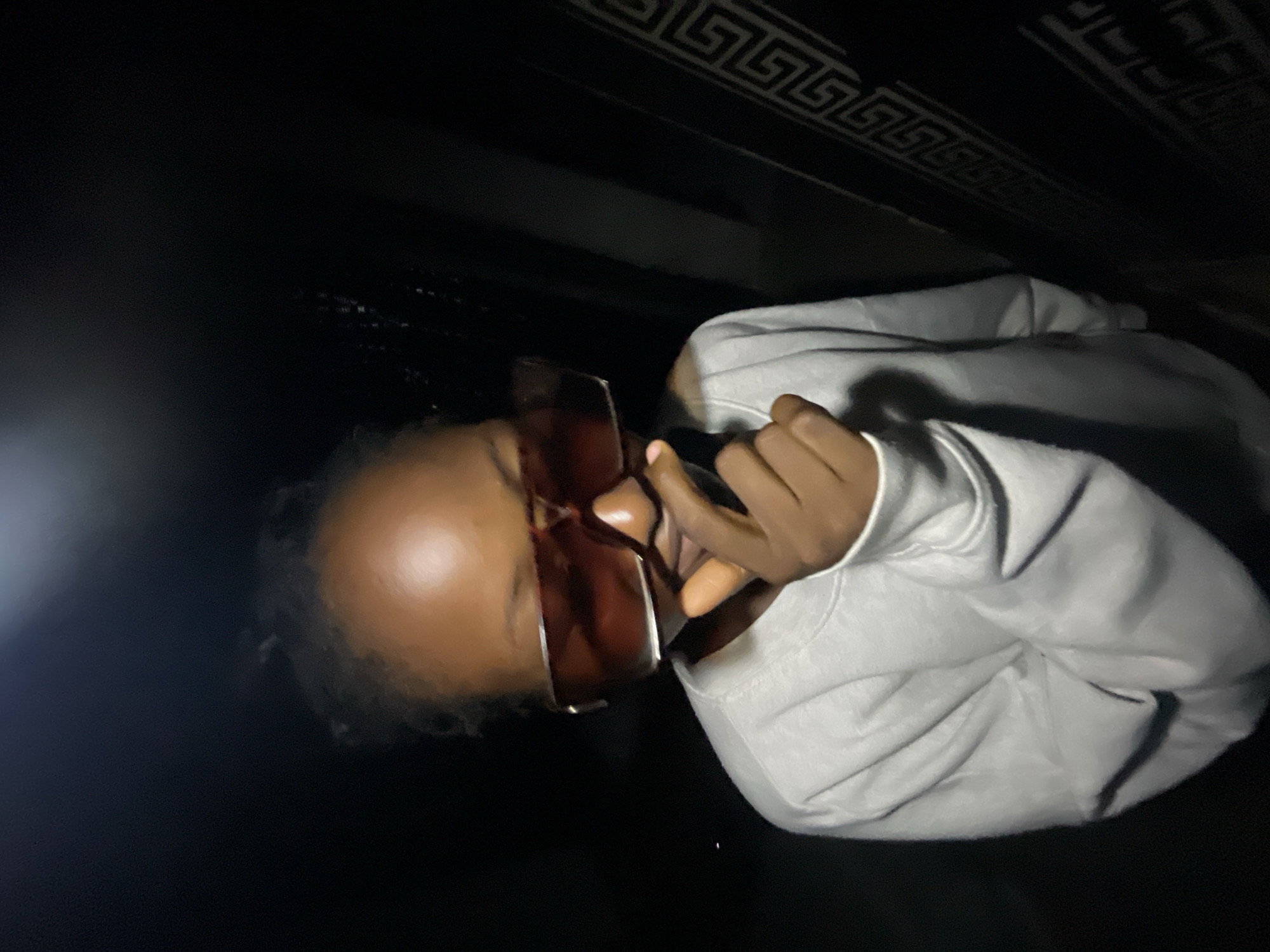 A medium shot captures a young Black individual wearing oversized, translucent brown sunglasses and a light gray crewneck sweatshirt. The subject is positioned in the foreground, with their right hand raised to their mouth, obscuring it as they look down. The lighting is dim, creating a dark environment, with a bright light source illuminating the subject's forehead and the left side of their face. To the right of the subject, a dark, patterned surface with a Greek key design is visible. The background is largely obscured by darkness, with some indistinct shapes suggesting an outdoor or dimly lit indoor environment. The scene is set in Lokoja, Nigeria, and appears to be a candid, nighttime photograph.