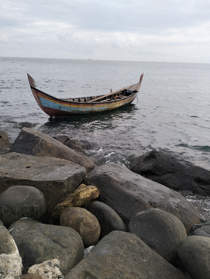 A weathered, colorful wooden boat rests on the water's surface, close to a rocky shoreline. The sky is overcast, casting a muted light over the scene. The boat displays a mix of red, blue, and yellow paint along its hull. Large, dark rocks fill the foreground, partially submerged in the water. The water appears calm, gently lapping against the rocks.