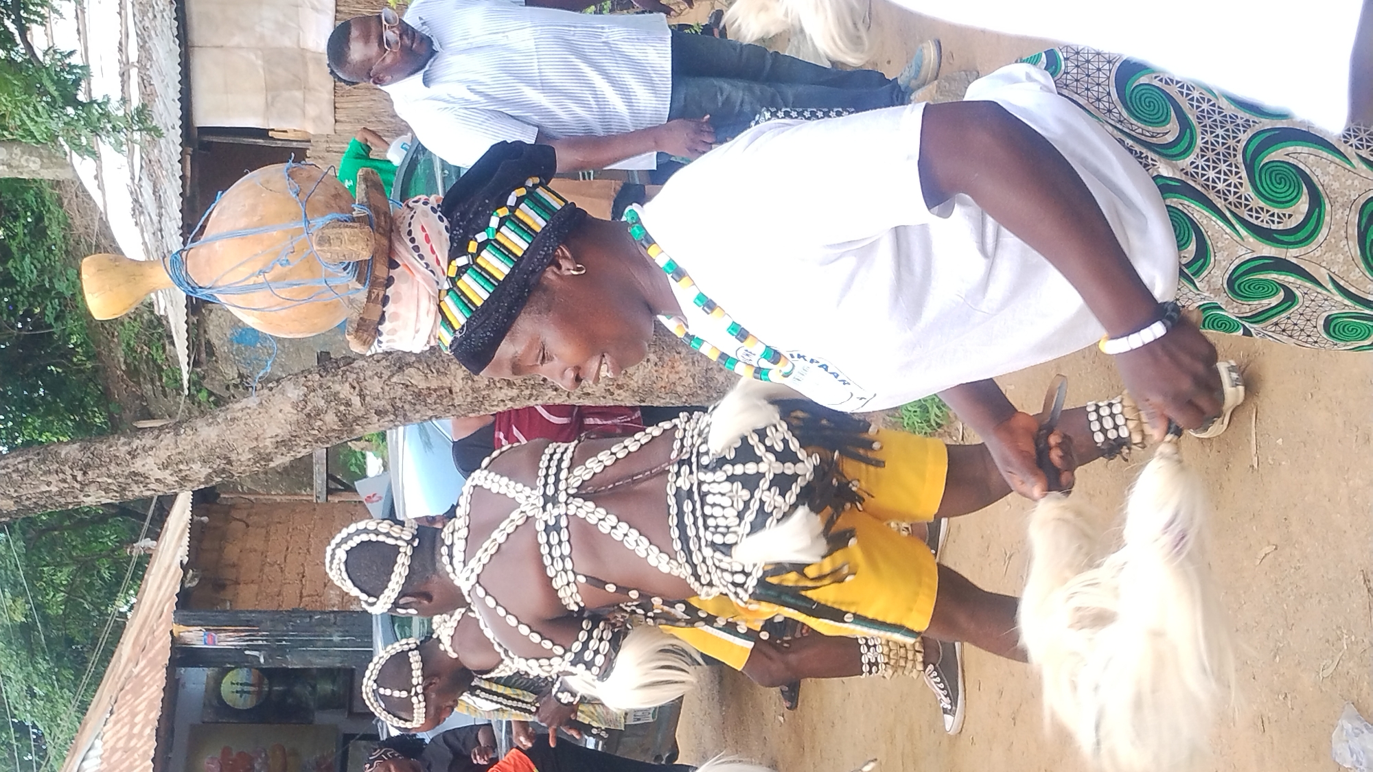 In the city of Jos, Nigeria, a group of people are gathered outdoors. The focus is on a woman in the foreground, wearing a white t-shirt and a colorful patterned skirt. She has a beaded headband and necklace. In her hands, she holds two white feathery objects, possibly for a dance or ritual. Behind her, two individuals are dressed in traditional attire adorned with cowrie shells and white feathers, with one wearing yellow shorts. They appear to be participating in a performance or ceremony. In the background, there are simple structures with corrugated iron roofs and mud walls, indicative of the local architecture. A man in a striped shirt and glasses stands to the right, observing the scene. The environment is dusty and appears to be a village or rural setting within Jos.
