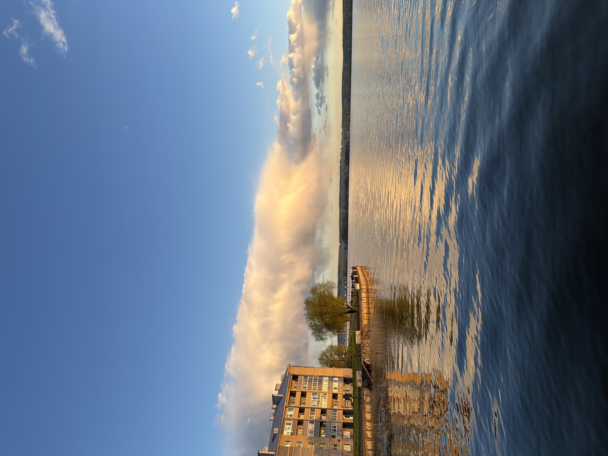 The image shows an outdoor scene in Votkinsk, Russia, on a body of water, likely a lake or river.
**Environment:** The sky is divided between a clear, light blue area and a massive bank of fluffy, illuminated clouds. The clouds appear to be catching the light of either sunrise or sunset. The water is slightly choppy, with reflections of the sky, clouds, and shore.
**Objects:** A multi-story building is situated on the shoreline, featuring numerous windows. Trees line the edge of the shore next to a fenced boardwalk.
**Details:** The reflections in the water mimic the colors and forms of the objects on the shoreline and in the sky. The overall scene conveys a sense of tranquility.