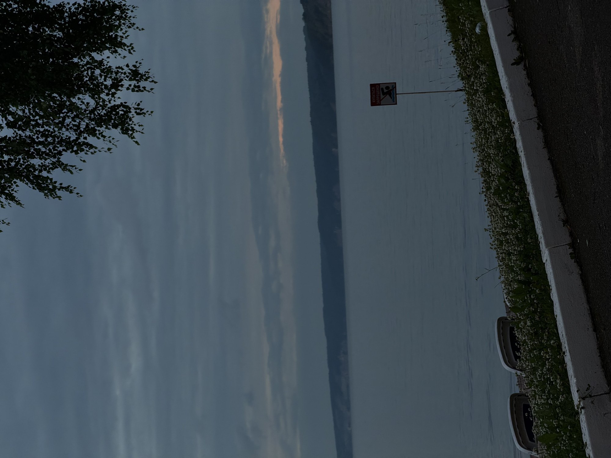 The image depicts a serene lakeside scene in Perm, Russia, likely during the late evening or early morning given the soft, muted light and overcast sky. A dark silhouette of trees frames the left side of the composition, contrasting with the expansive, calm water that dominates the middle ground. In the distance, a long, elevated landmass, possibly a cliff or a series of hills, stretches across the horizon.
Along the right bank, a white concrete curb separates the water from a paved area. This bank is densely lined with small white flowers and green foliage, creating a vibrant border against the muted tones of the water and sky. Two oval-shaped structures, possibly planters or decorative elements, are visible amidst the flowers.
A prominent red and white sign stands near the water's edge. The text on the sign, though partially obscured and oriented vertically, appears to be in Cyrillic script and depicts a silhouette of a person diving into water with a diagonal red line through it, indicating a prohibition against diving. This suggests the area may be a public space near a body of water.
The overall mood is tranquil and somewhat somber, with the low light and subdued colors contributing to a peaceful, contemplative atmosphere. The gentle ripples on the water and the distant landscape create a sense of depth and scale. The scene evokes a feeling of quietude and natural beauty.