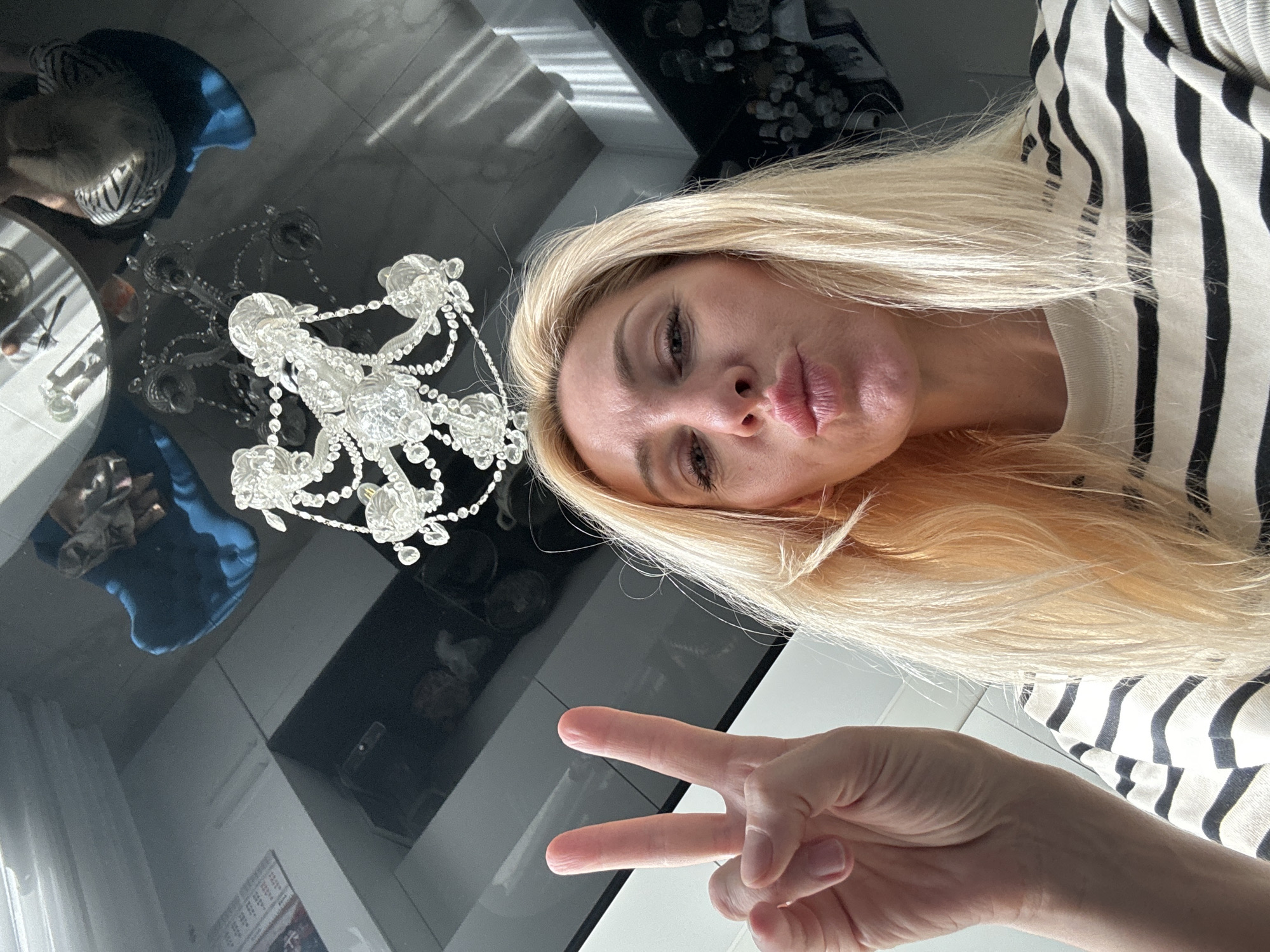 The image is a close-up selfie taken from a low angle, looking upwards at a woman. She has blonde hair and is making a kissing face. Her right hand is raised, displaying a peace sign. She is wearing a black and white striped shirt. The background appears to be a modern interior with a glossy black ceiling reflecting a chandelier and furniture. There is no indication of the city of Lednevo, Russia in the image.