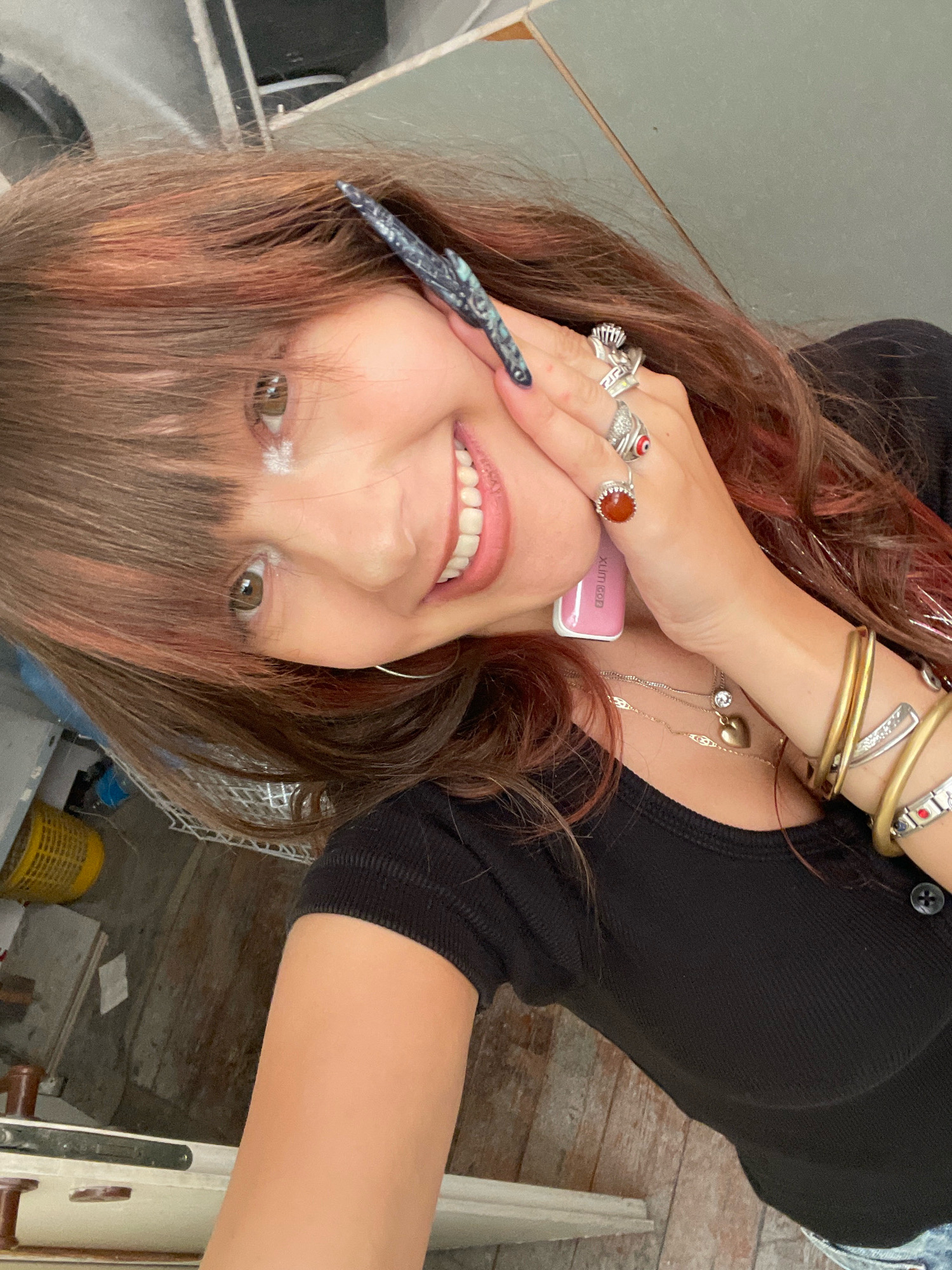 A woman with brown hair with red undertones and bangs smiles at the camera. She is wearing a black ribbed t-shirt and has several rings on her fingers, as well as bracelets on her wrist. She is holding a pink electronic device and has a light dusting of white powder on her eyelids. The background appears to be an interior space with worn wooden flooring and exposed shelving. The image was taken in Raanana, Israel.