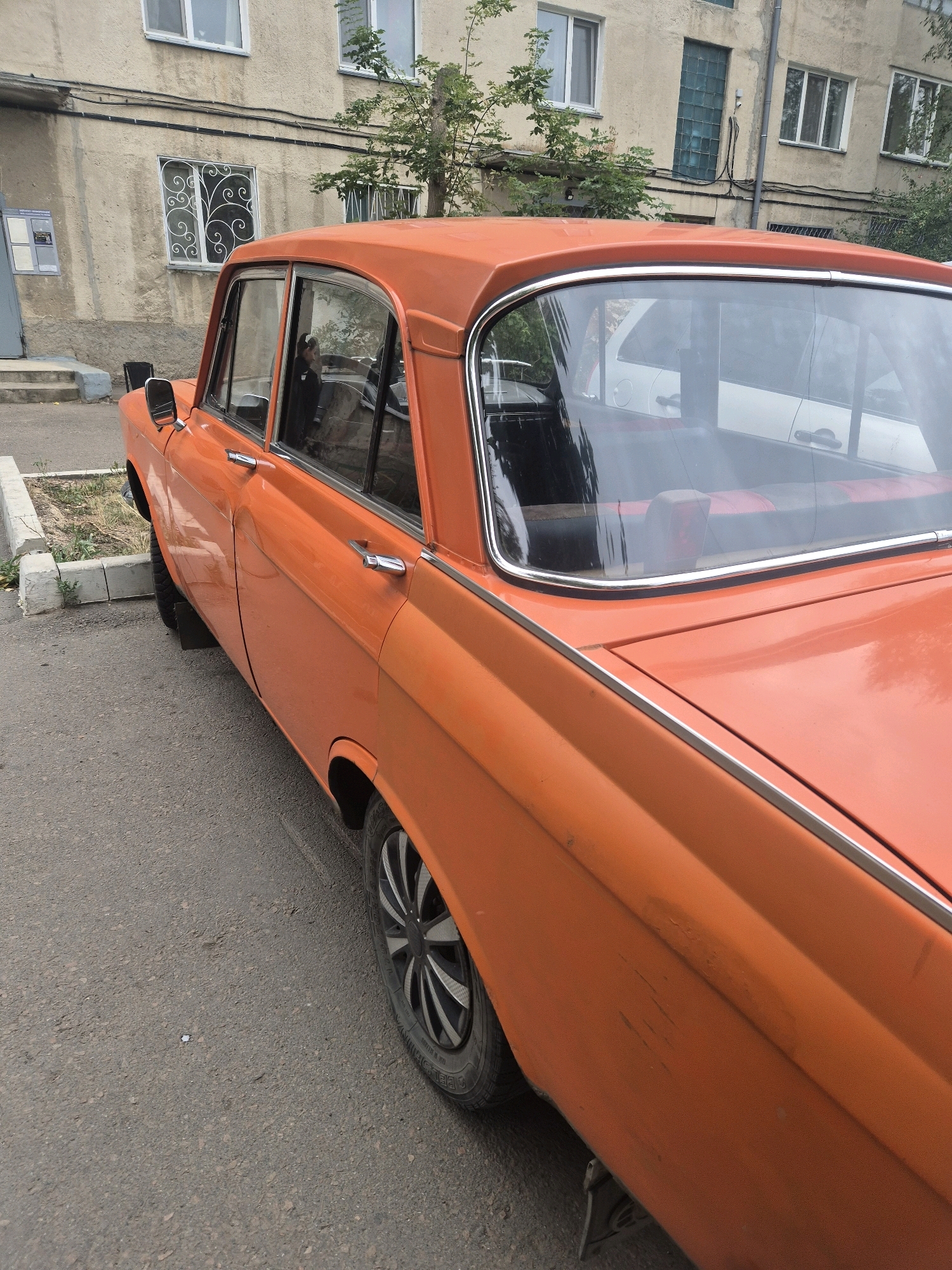 The image captures a close-up, angled view of the rear passenger side of an orange sedan parked on an asphalt road. The car has a chrome trim line running along its side and a distinctive chrome strip framing the rear window. The window itself reveals a partially visible interior with red and black striped seats and what appears to be a person reflected in the glass. In the background, a light-colored, multi-story apartment building with visible windows and some greenery suggests an urban residential setting. The building's facade is textured and appears weathered, with electrical conduits running horizontally. To the left of the car, a concrete curb delineates a patch of dry grass and dirt. The overall scene is mundane and appears to be a typical street view in a city. This specific scene is located on Abylai Khan Avenue in Kokshetau, Kazakhstan, identified by the distinctive architecture and urban layout consistent with the area.