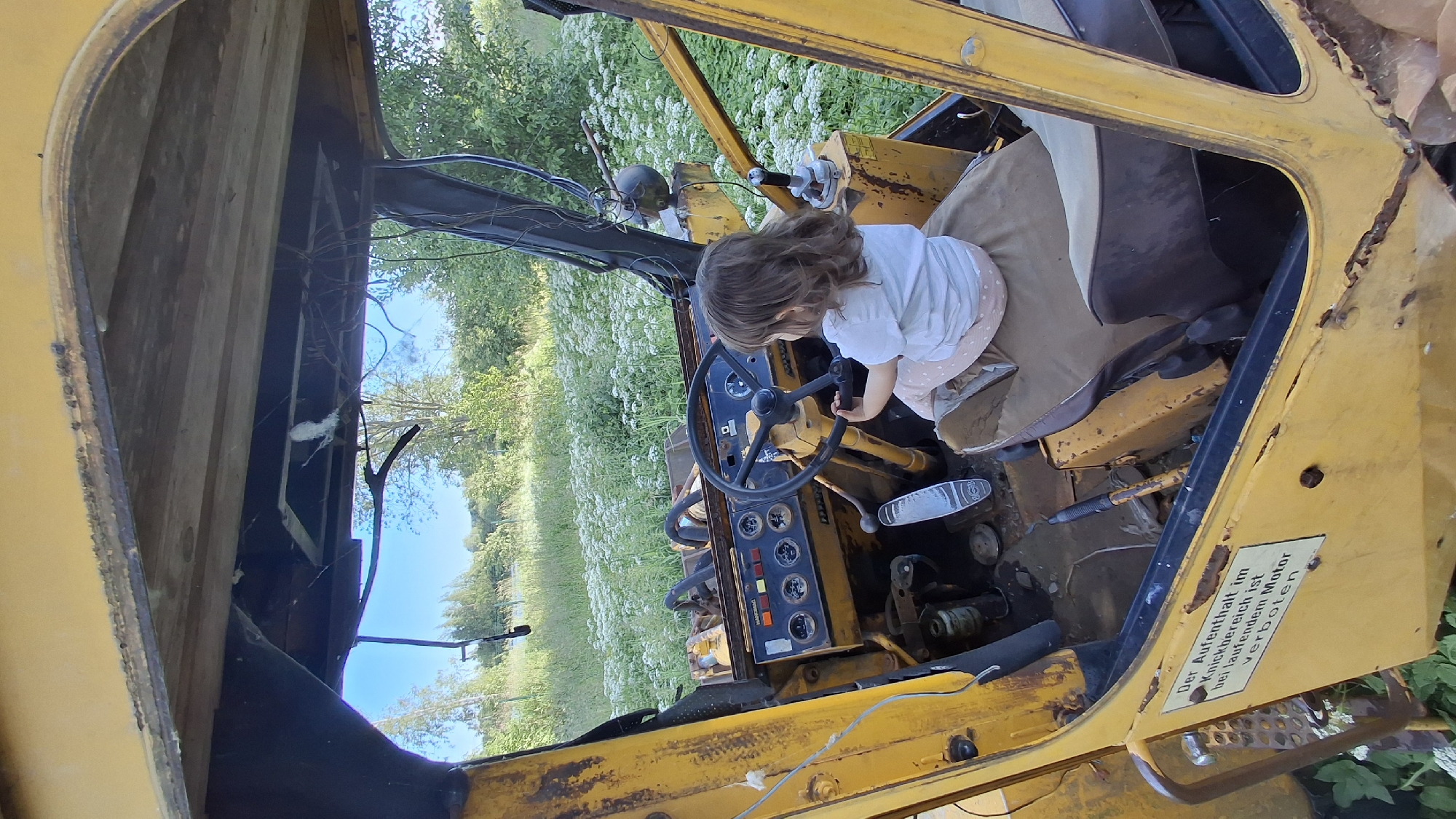 A young child with light brown hair is sitting in the driver's seat of a yellow, rusty, possibly abandoned vehicle, likely a tractor or construction equipment. The child is wearing a white t-shirt and light-colored pants with a polka dot pattern. They are holding onto the steering wheel, appearing to be playing or exploring the interior of the vehicle.
The scene is set outdoors in a grassy field with abundant white wildflowers and green foliage. The sun is bright, suggesting it is daytime, and the weather appears to be fair. The vehicle's interior is cluttered with wires and mechanical parts, indicating it may not be in working condition. A seat covered in a tan and brown fabric is visible behind the child.
A yellow sticker with German text is affixed to the exterior of the vehicle, near the door. The text reads: "Der Aufenthalt im Knickbereich ist bei laufendem Motor verboten." This translates to: "Staying in the bending area is forbidden when the engine is running."
The overall impression is one of a child's innocent exploration in a somewhat neglected environment. The strong yellow of the vehicle contrasts with the natural green and white of the surroundings.