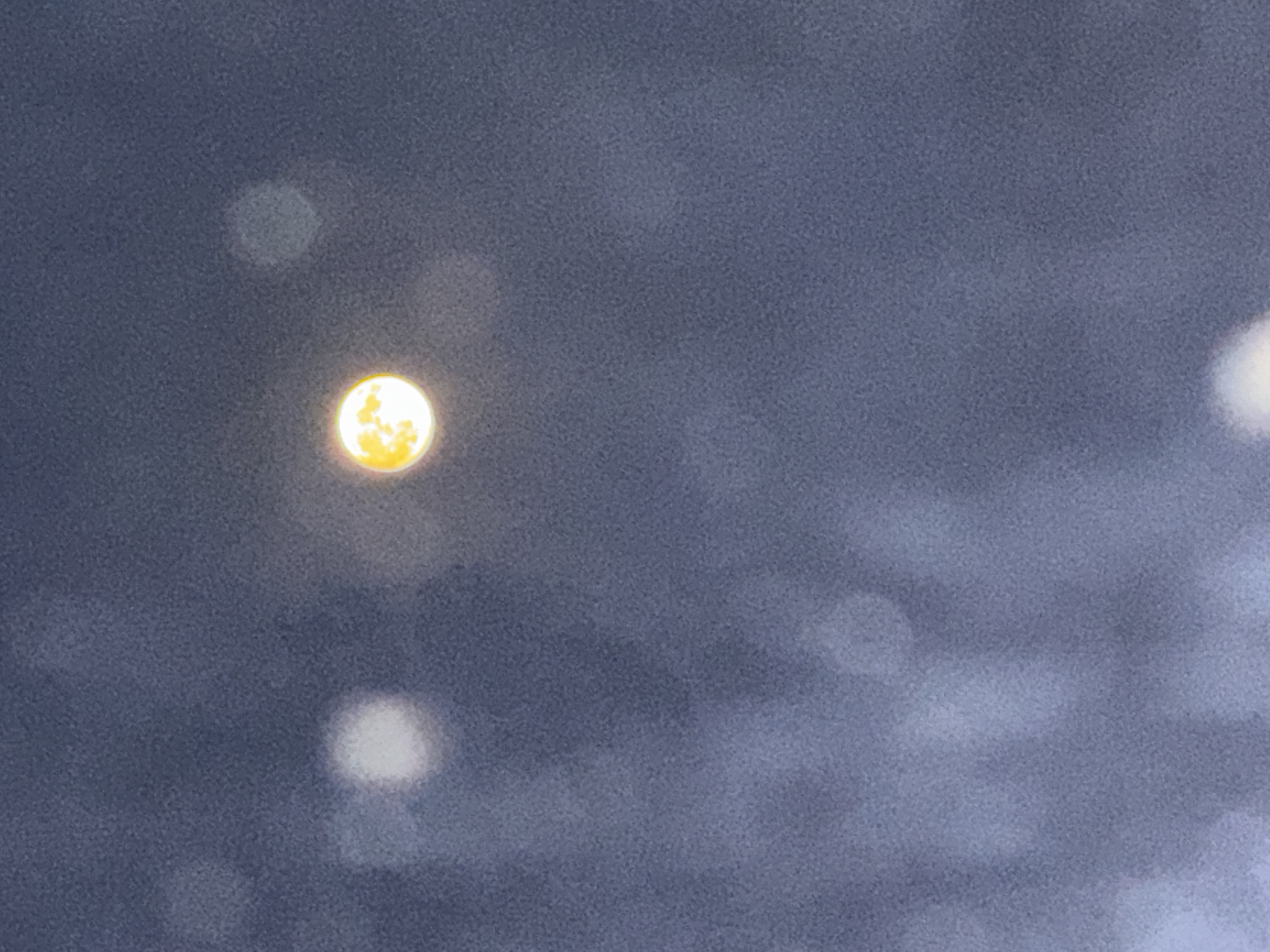 The image shows a bright, yellow-orange moon against a dark, grainy sky. There are several out-of-focus circular shapes, likely representing lights or bokeh effects, scattered across the frame, primarily in the upper and lower portions. The overall impression is of a night scene, possibly taken with a phone camera with some motion blur or atmospheric conditions affecting the clarity. There is no discernible indication of the city of Zatoka, Ukraine, or any specific landmarks within the image itself.