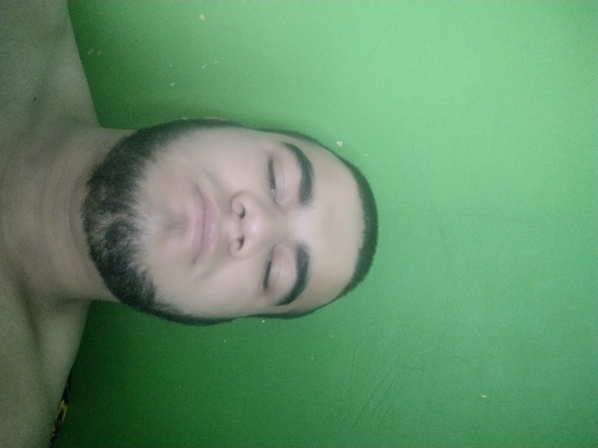 The media file features a close-up, sideways shot of a young person lying down against a solid green background. The subject has dark hair and a beard, with their eyes closed and their mouth slightly pursed. Their facial expression appears relaxed or perhaps sleepy. The lighting is somewhat dim, casting soft shadows on their face. The background is a plain, vibrant green, possibly a wall or fabric backdrop. A few small white specks are visible on the green surface. The overall impression is a candid, informal portrait, possibly taken in a casual indoor setting. Given the provided location context of "казарма" (barracks), it is plausible that the scene takes place within a military barracks environment in Russia. No text is visible in the image.