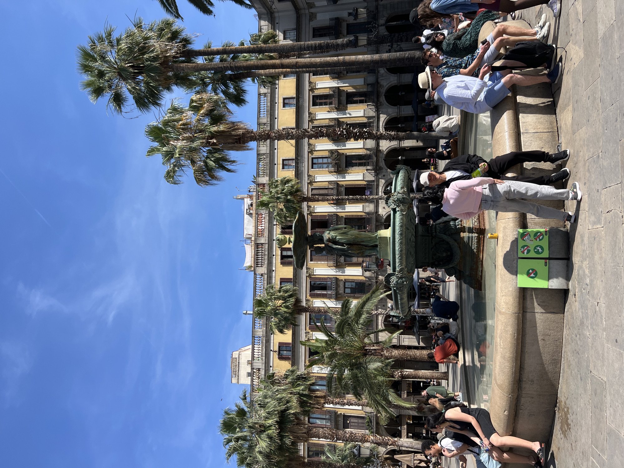 The image depicts a sunny scene in Barcelona, Spain, likely in Plaça de Catalunya. The foreground features a stone fountain with people sitting along its edge. Palm trees are interspersed throughout the square. Behind the fountain, there is a multi-story building with yellow walls and balconies. A clear blue sky is visible above. People are seen sitting, standing, and interacting around the fountain. The ground appears to be paved with light-colored stone. Two green bins are seen at the edge of the fountain near the right.