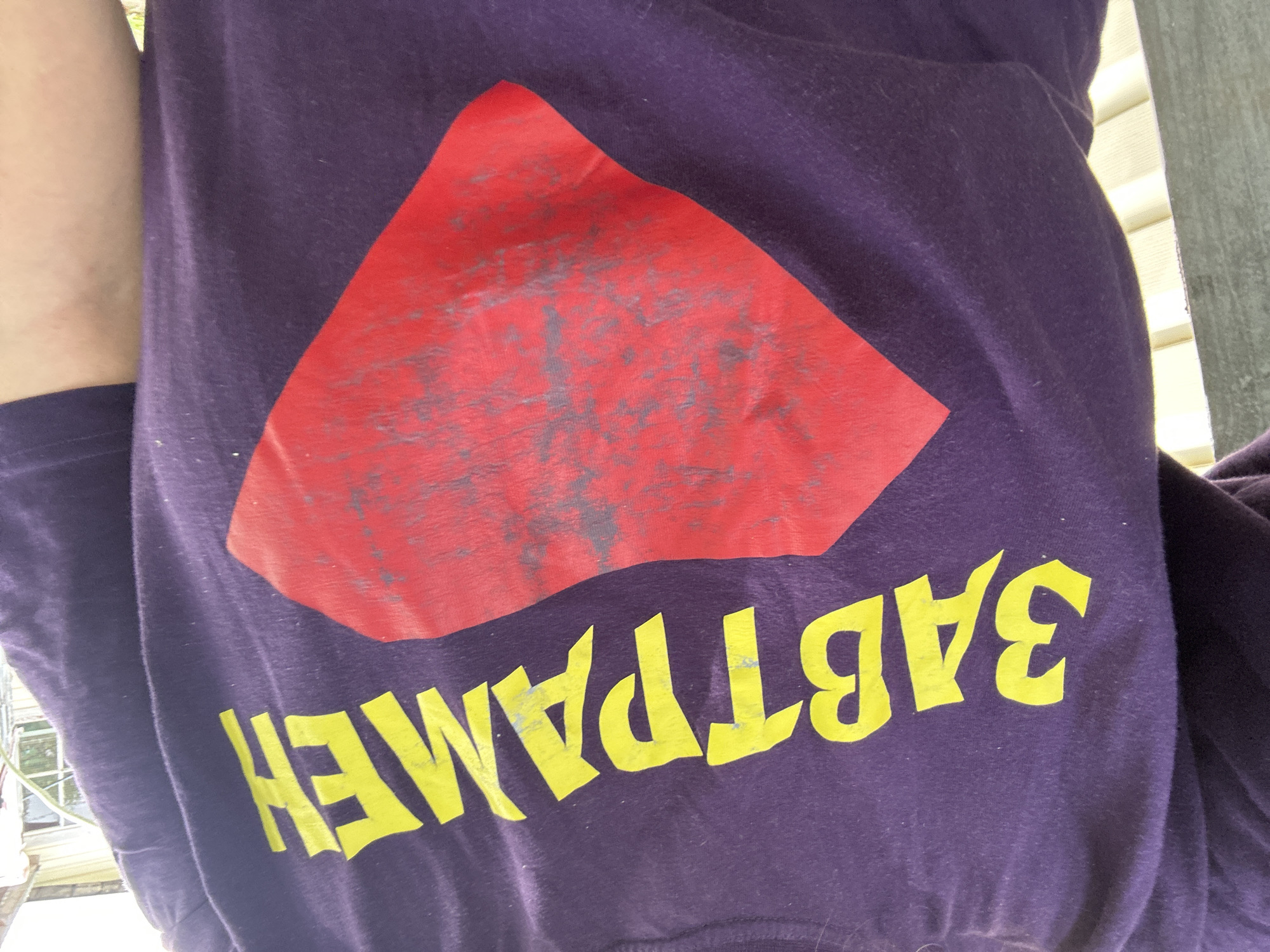 A person wearing a purple t-shirt with a yellow Cyrillic text and a red diamond-shaped emblem is pictured outdoors. The text on the t-shirt appears to read "ЗАВТРАМЕН" (Zavtramen). The red emblem has a distressed or faded appearance with greyish markings. The background shows siding of a building, possibly a house, and a white railing or fence. The lighting suggests it is daytime. The scene is not specifically located in the city of Ленинградская, Russia.
