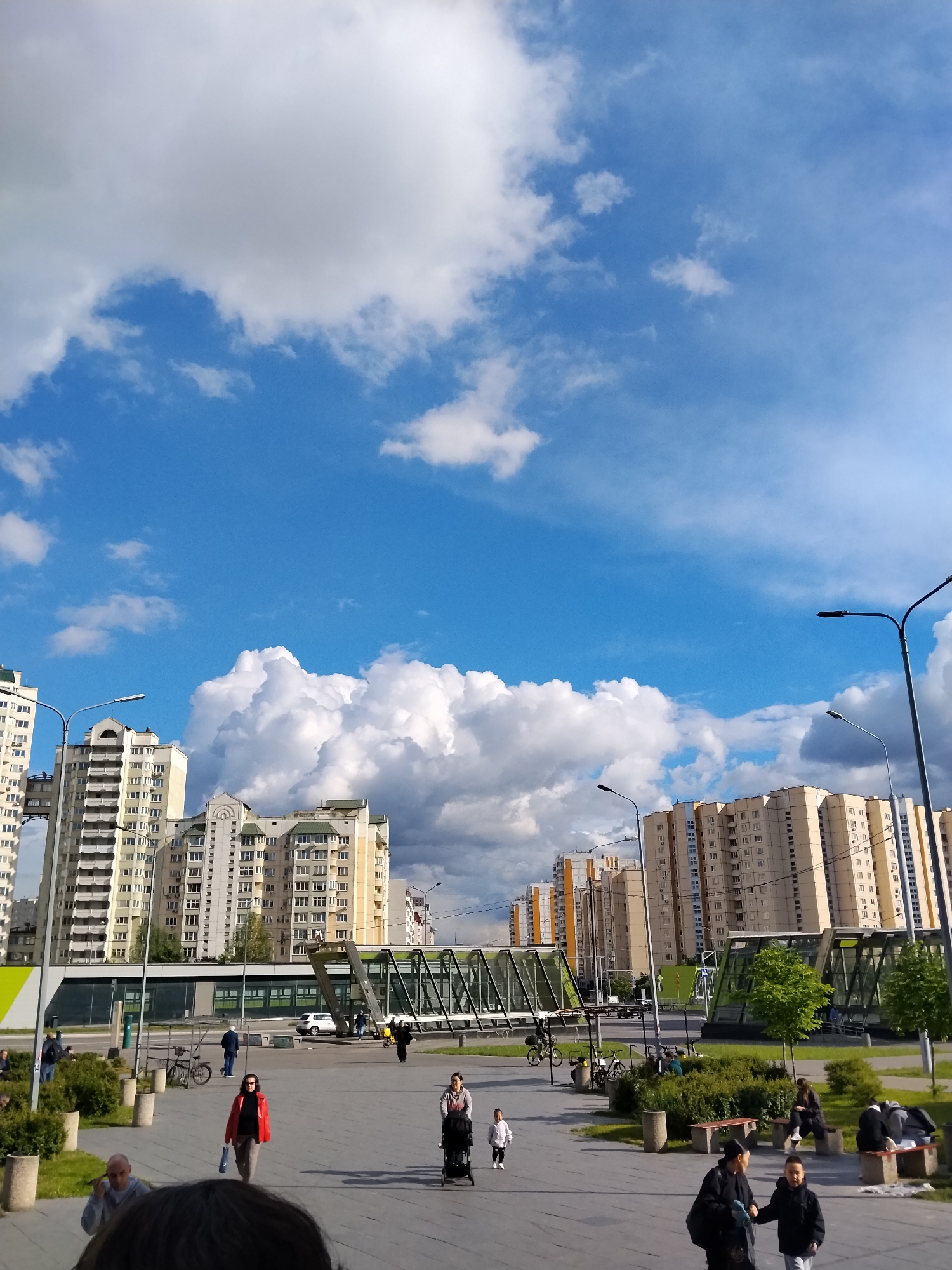 The image captures an urban scene under a sky with vibrant blue patches interspersed with puffy white clouds. In the middle ground, several high-rise apartment buildings of beige and off-white colors line the street. A pedestrian bridge spans over a thoroughfare, its entrance highlighted by green glass structures on either side. Pedestrians populate the walkway, including a woman in a red coat, a mother pushing a stroller, and individuals sitting on benches. Street lamps cast shadows along the paved area. The scene is located in the city of Unknown, Unknown.