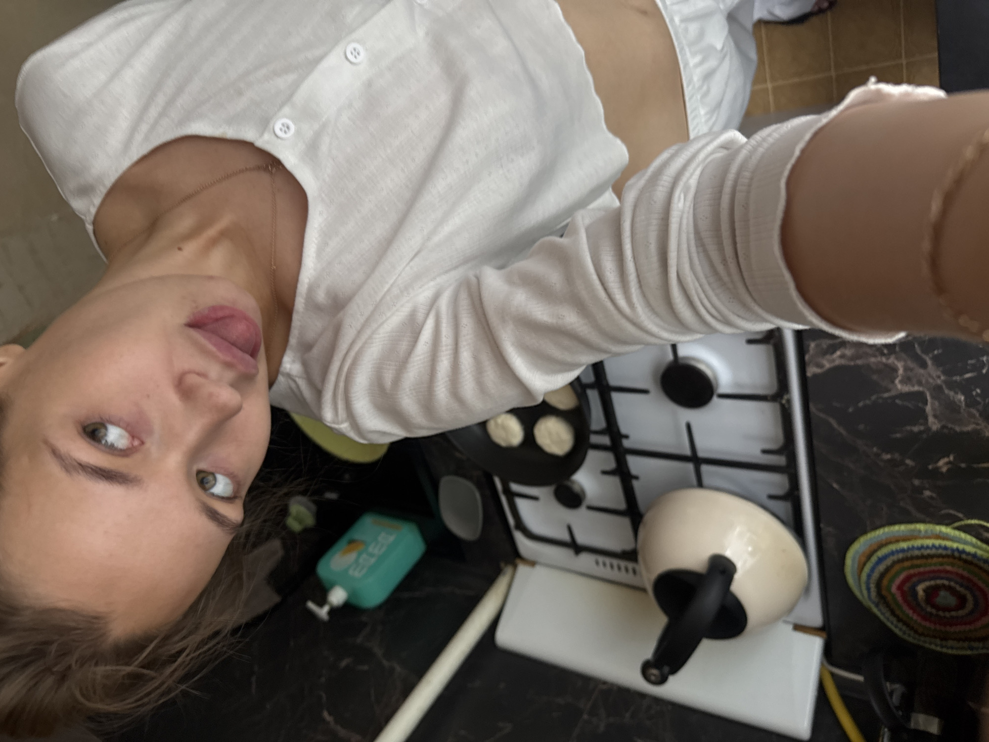 The media file shows a close-up, self-taken photograph of a young woman in a kitchen. She is wearing a white, button-up shirt with a textured pattern and a long-sleeved, ribbed white cardigan. The woman is looking directly at the camera, with her tongue sticking out slightly. Her eyes are wide open and appear to be a light green color. Her blonde hair is slightly messy and falls around her face.
In the background, a white gas stove is visible with a small frying pan containing several round batter or dough items, possibly pancakes or fritters. A cream-colored teapot with a black handle rests on the stovetop. To the left of the stove, there is a turquoise bottle of liquid soap with a white pump dispenser. The countertop is dark and appears to be made of marble or a similar material. To the right of the teapot, a circular woven mat with colorful concentric rings is visible. The overall lighting suggests it is daytime, possibly indoors with artificial light. The woman's expression is playful and relaxed. The context provided indicates the location is Chaikovsky, Russia.