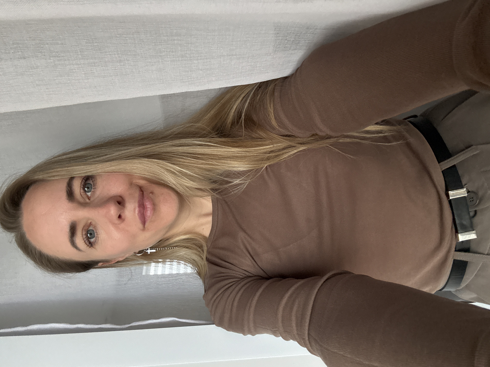 A selfie taken indoors in Chaikovsky, Russia, features a young to middle-aged woman looking directly into the camera. The woman has long, straight blonde hair that falls around her shoulders. Her face displays clear skin, light makeup, blue-grey eyes, and a gentle, closed-mouth smile.
She is wearing a brown, long-sleeved top with a subtle ribbed texture. A black belt with two distinct silver rectangular buckles is visible at her waist, suggesting she is wearing high-waisted trousers or a skirt in a dark, possibly olive or grey, color. A silver earring shaped like a cross hangs from her left ear.
The background is simple, consisting of a light-colored, possibly white or light grey, fabric curtain or blind, indicating an indoor setting likely near a window. The lighting appears soft and natural, suggesting it is daytime. The woman's expression is calm and composed. No visible text is present in the image.