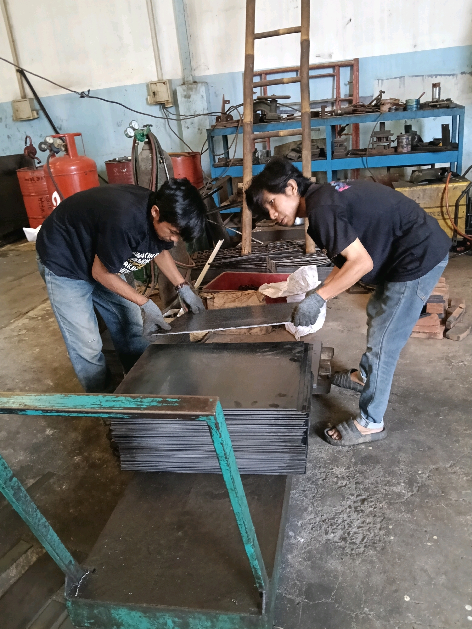 In a workshop environment, two men in black t-shirts and blue jeans are actively engaged in handling a stack of large, flat metal sheets. One man, on the left, is bent over, his hands wearing grey gloves, as he appears to be separating or moving a sheet from the top of the pile. The other man, on the right, is also wearing grey gloves and is in a similar bent posture, reaching towards the metal sheets. He is wearing dark-colored sandals. A large stack of metal sheets is prominently displayed on a green metal cart or platform.
The workshop setting includes various industrial elements. To the left, a red gas cylinder with gauges is visible, alongside other metallic barrels and equipment. In the background, a tall wooden ladder leans against a blue metal shelving unit filled with tools and equipment. The walls are painted a light blue, and the floor is a concrete surface, showing signs of wear and some debris. The overall impression is of an active industrial workspace in Belimbing, Indonesia, where metal fabrication or processing is taking place.