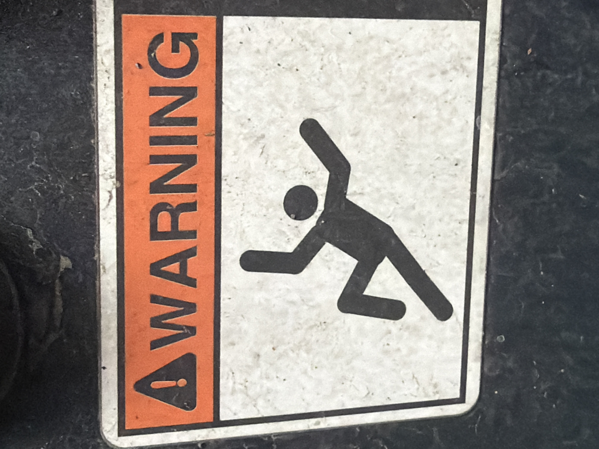 A warning sign is affixed to a dark, textured surface. The sign has an orange header with a black exclamation point inside a triangle and the word "WARNING" in black block letters. Below the header, a black pictogram depicts a person falling. The pictogram shows a figure with arms and legs outstretched as if in mid-fall. The background of the pictogram area is a mottled white. The sign appears to be made of metal or a durable plastic and shows signs of wear, including some scuffs and dirt. The surrounding dark surface appears to be part of a piece of machinery or equipment. This image was taken in Murfreesboro, United States of America.