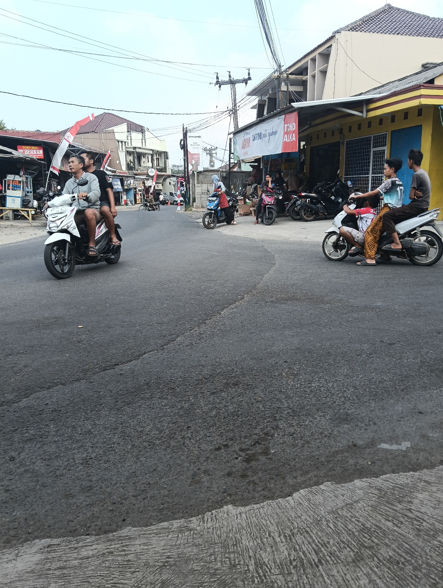 In Puspahiang, Indonesia, this image captures a street scene with several motorbikes. In the foreground, two individuals are riding on a white motorbike. The driver, a man with graying hair, wears a gray t-shirt and shorts. The passenger behind him wears a black t-shirt and shorts. Further down the road, another motorbike carries two young people. The driver, a male in a patterned shirt and shorts, is followed by a male passenger in a dark t-shirt and pants. A third motorbike is visible with two passengers, one wearing a red and white patterned top and the other a blue t-shirt. On the left side of the street, a shop with a red and white flag is visible, and several other motorbikes are parked. The road surface is asphalt, with a concrete shoulder. Buildings line the right side of the street, including a yellow building with a blue door and a building with a white banner. Overhead, electrical wires are strung across the sky. The sky is overcast.