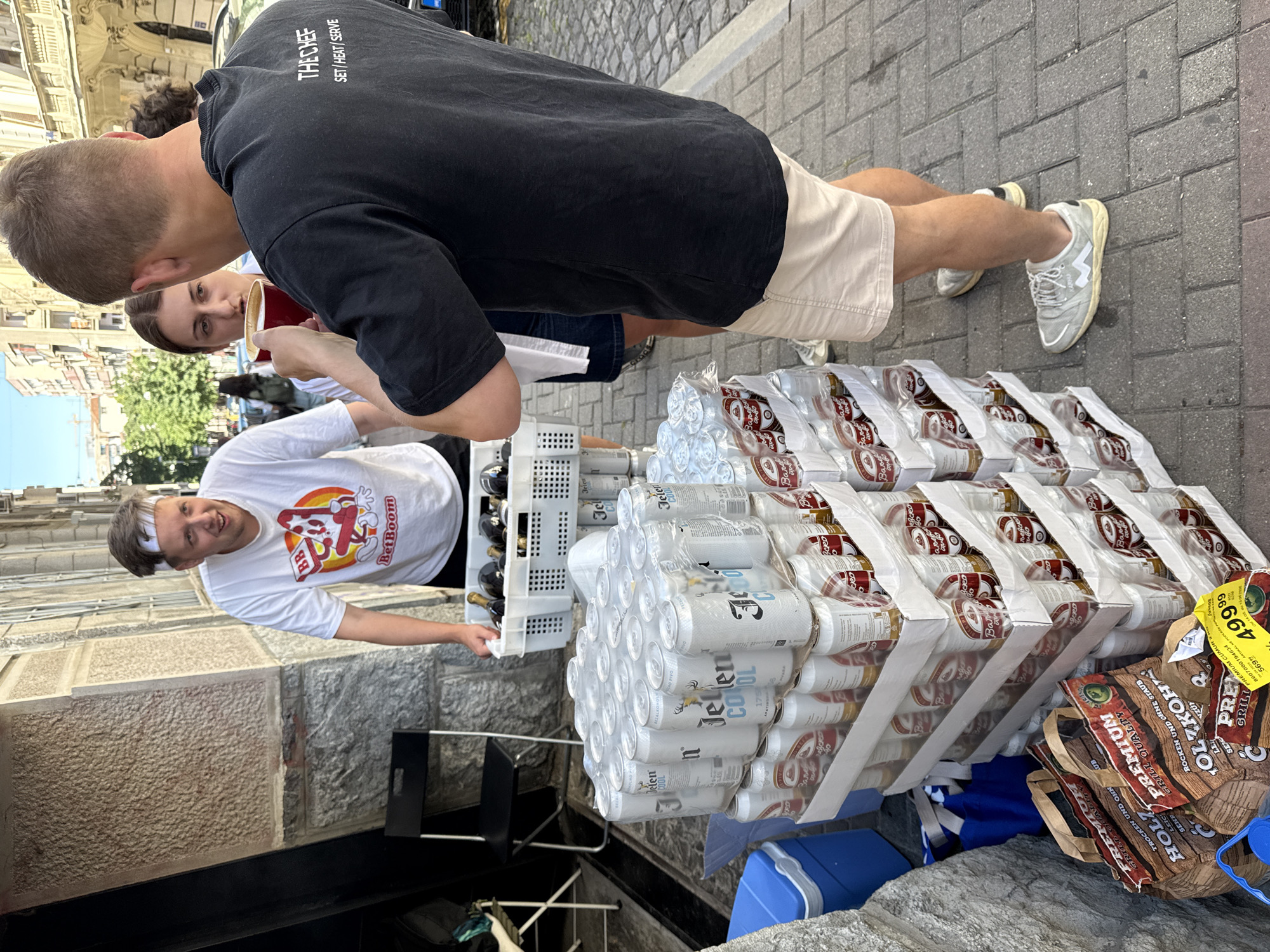 The image shows a street scene in Belgrade, Serbia. A Caucasian male wearing a white t-shirt with a "BetBoom" logo is holding a plastic crate with bottles. Adjacent to him is a tower of canned "Jelen COOL" beer and bagged "PREMIUM GRILL" wood. To the right is a man wearing a black "THE CHEF" t-shirt and beige shorts. Another person is behind him, holding a red cup. The setting is outdoors on a brick-paved street. The environment includes stone walls and other urban elements.