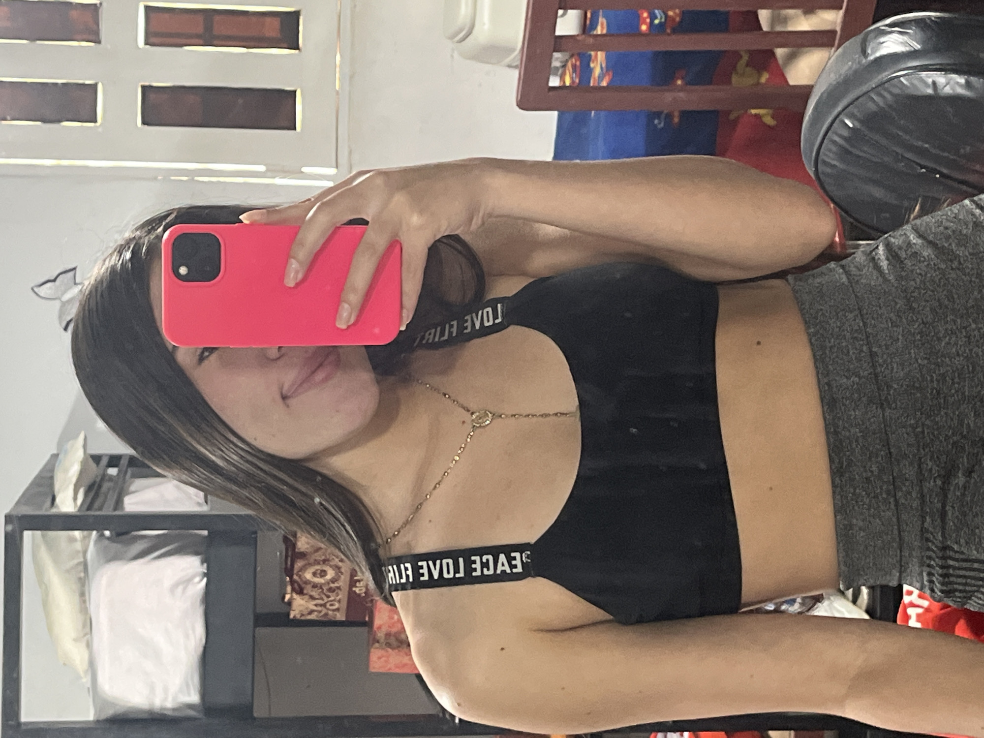 A young woman is captured in a mirror selfie, holding a bright pink smartphone that partially covers her face. She has long, dark, straight hair and displays a subtle smile.
She is wearing a black sports bra with a white strap that features the reversed text "PEACE LOVE ELIF." Below this, she wears gray, textured athletic shorts or leggings. A delicate gold body chain is draped across her chest and torso.
The background, visible in the mirror's reflection, suggests an indoor, casual setting, likely a home in Juan Griego, Venezuela. To the left of the reflection (the woman's right), a white wall with four rectangular window-like openings, possibly with dark panes, is visible. Below these, a dark, open shelf holds several white, soft items, potentially bags or folded materials. To the right of the reflection (the woman's left), parts of a wooden furniture piece, possibly a chair or table, with blue and red patterned fabric, can be seen. Adjacent to this, a dark, rounded object, possibly a leather cushion or small punching bag, is present.
The image is well-lit, providing clear details of her attire and the surrounding environment. The visible text on her sports bra strap reads "PEACE LOVE ELIF" when correctly oriented.