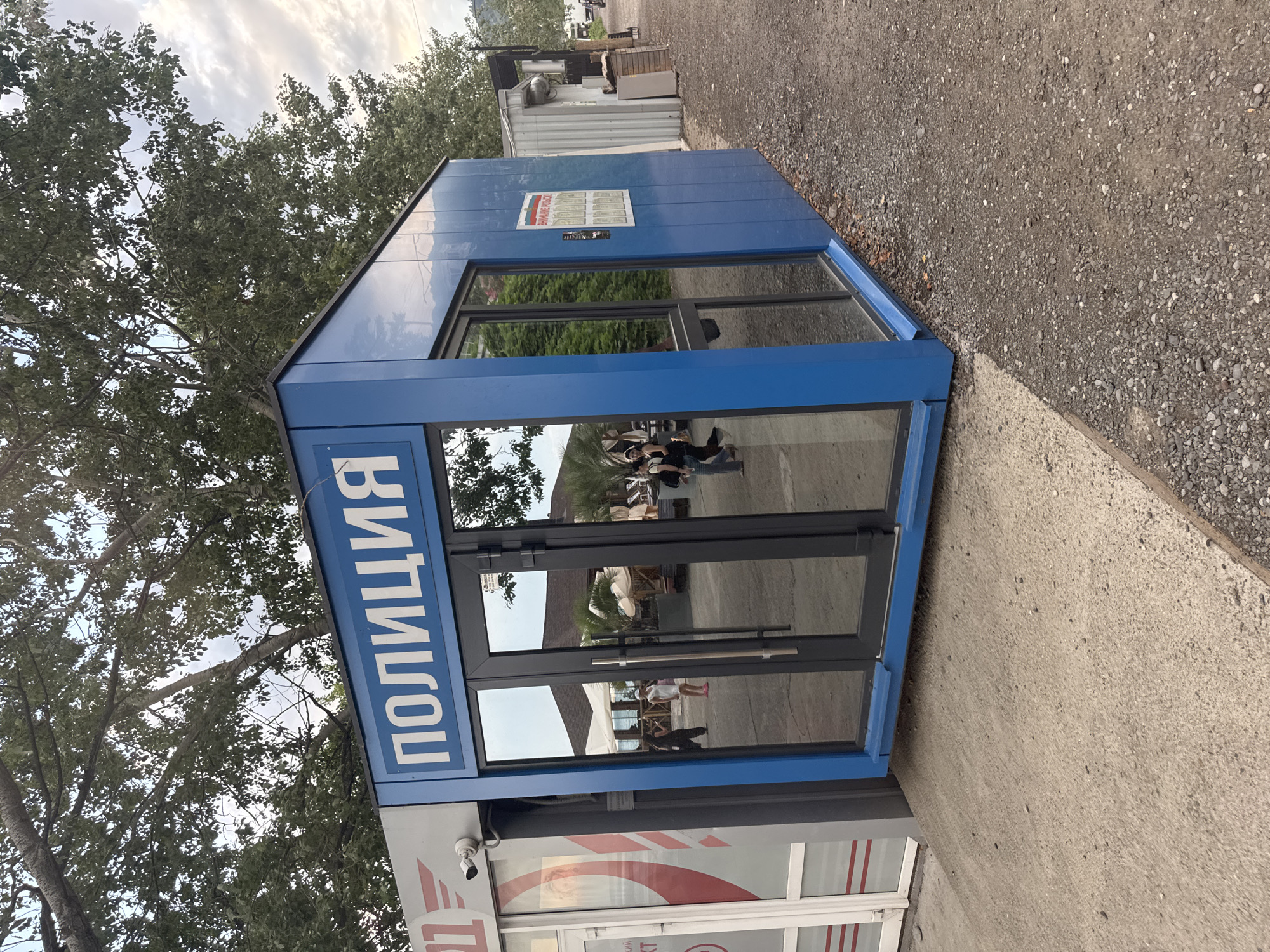 A blue police kiosk is situated on a gravel and concrete path next to other small structures and trees. The kiosk has large glass doors and windows reflecting the surroundings. The word "ПОЛИЦИЯ" (Police) is written in large white letters on the front of the kiosk. Several people are visible in the reflection of the glass doors, walking and standing. In the background, there are more buildings, trees, and a cloudy sky. The scene appears to be an outdoor public space, possibly near a beach or resort area.