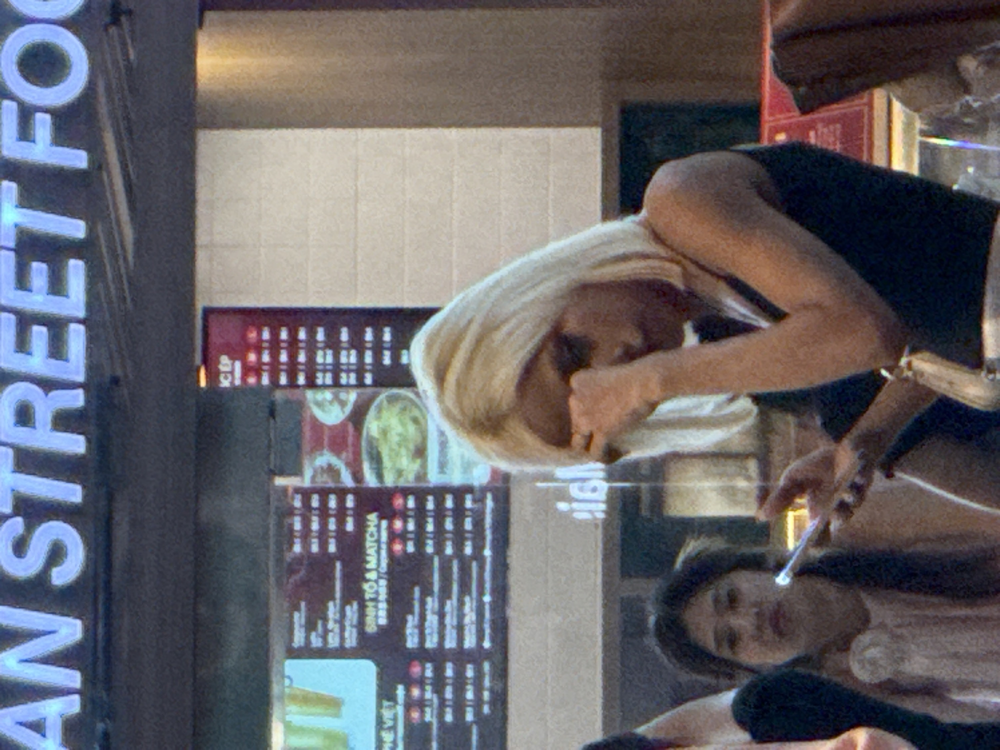 The image shows a street food stall, likely in Bắc Cam Ranh, Vietnam. In the foreground, a person with short blonde hair is holding a phone and appears to be rubbing their eye. Behind them, another person with dark hair is visible, also holding a phone. The stall features a menu board with various food and drink items. The overall lighting suggests an indoor or evening setting.