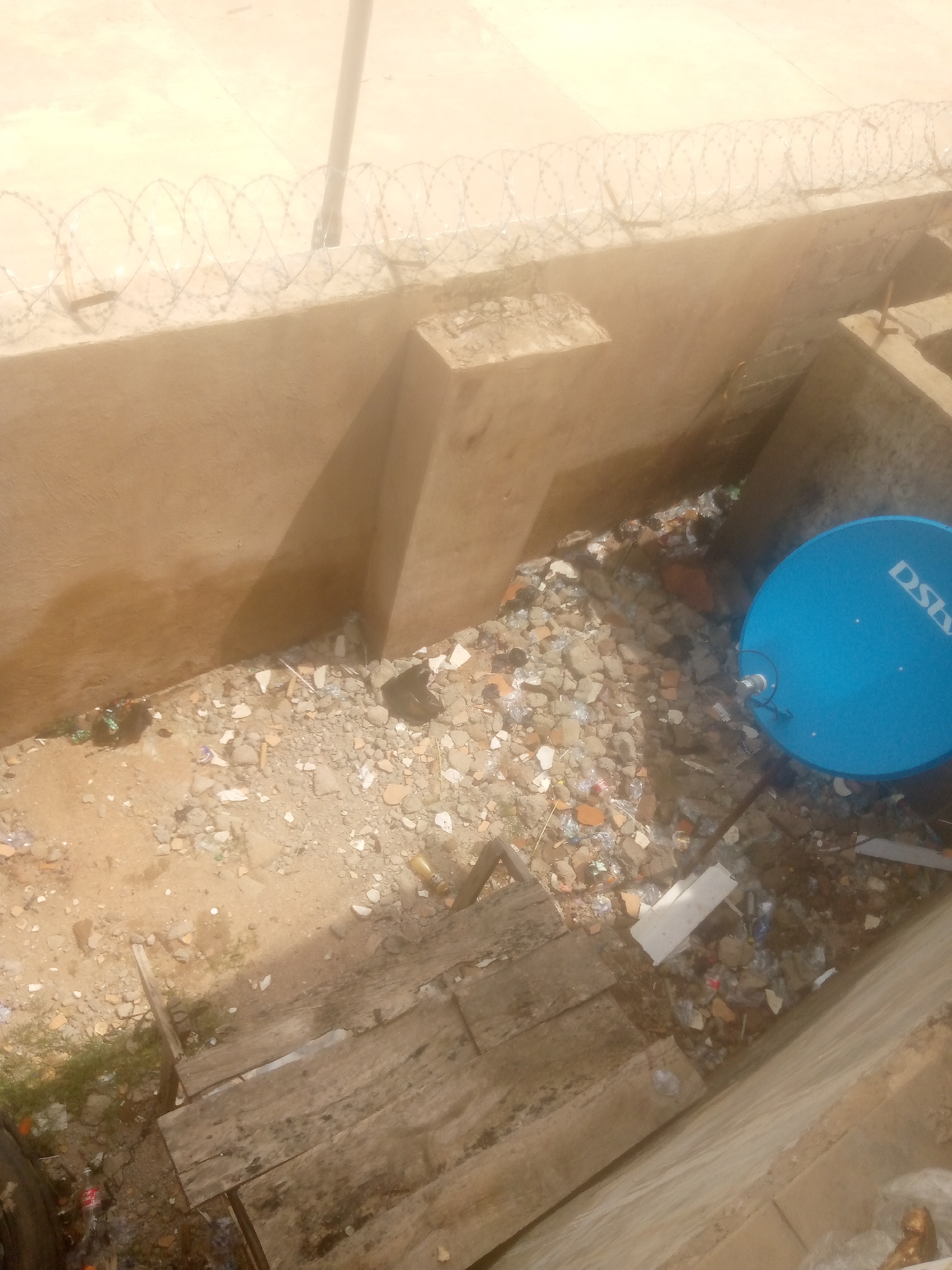 The image depicts a narrow, outdoor alleyway or drainage channel filled with a significant amount of debris. In the upper portion of the image, a concrete wall is topped with barbed wire fencing. Below this, the channel floor is uneven and covered in scattered rubble, including broken bricks, plastic fragments, and various other pieces of trash. A large blue satellite dish, possibly for DSTV, is visible on the right side of the channel. To the left, a section of wooden planks is laid out. The overall impression is one of neglect and disrepair. The scene appears to be in an urban environment, possibly a low-income area or a less maintained section of Jalingo, Nigeria.