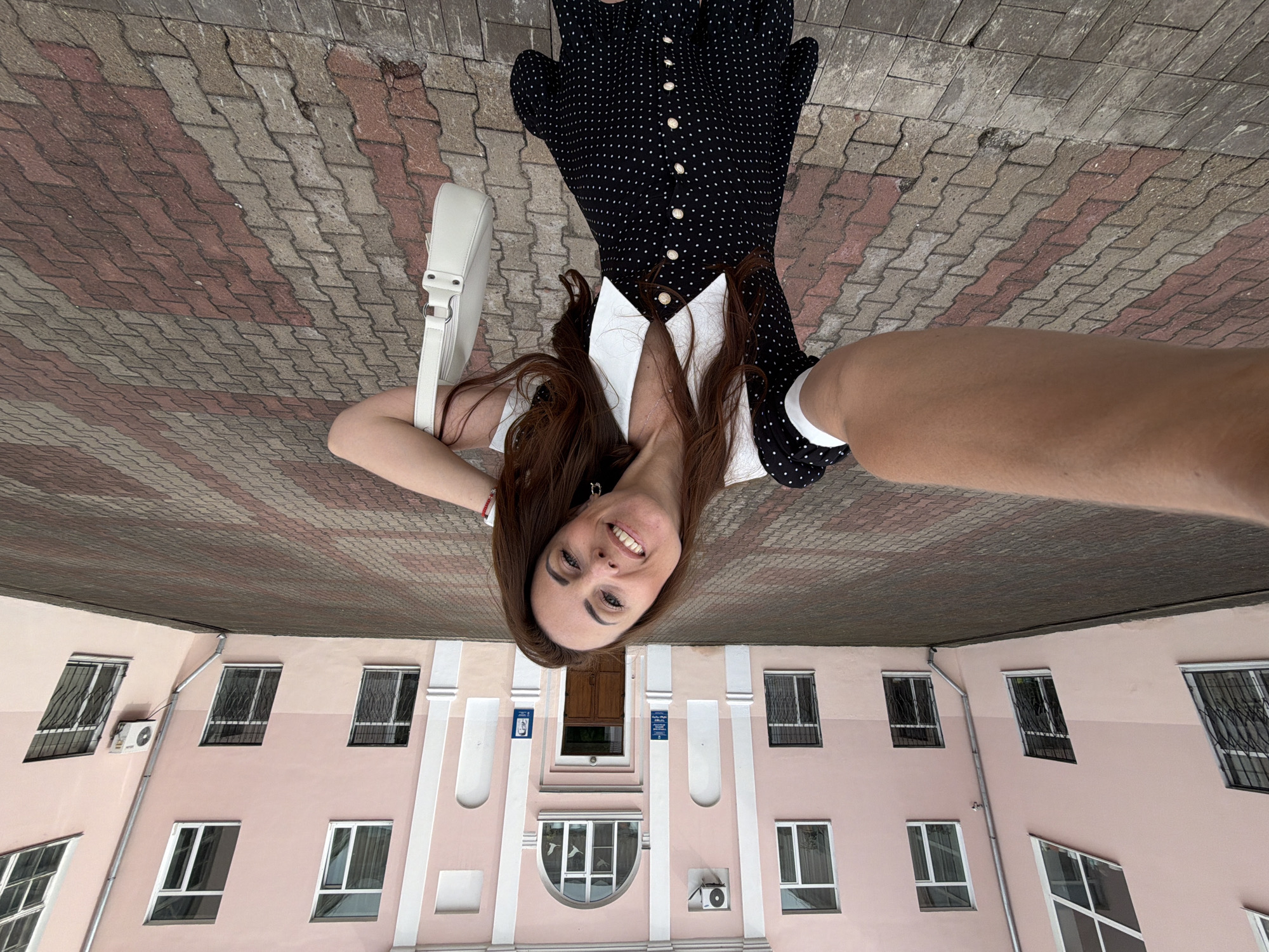 A woman is taking a selfie in front of a pink building. The building has multiple windows and an arched entrance. The woman is wearing a black and white polka dot dress and holding a white purse. The ground is paved with interlocking bricks in a pattern of red and grey. The scene appears to be a public space, possibly a school or administrative building, located in the city of Birobidzhan, Russia.