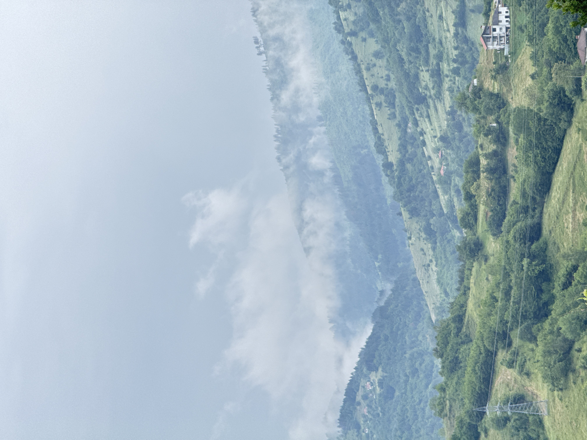 This detailed image depicts a vast, verdant mountainous landscape situated near Ardino, Bulgaria, under a heavily overcast sky. The upper third of the frame shows a pale blue-grey sky, with diffuse white and light grey clouds or fog partially obscuring the mid-ground mountain peaks. Multiple successive layers of mountains, covered in dense green deciduous and coniferous forests and lighter green grassy slopes, recede into the distance, creating significant atmospheric perspective. In the lower-left quadrant, a large, grey metal electricity transmission tower stands prominently on a grassy incline, with power lines extending horizontally through the middle ground. In the lower-right quadrant, a multi-story white house with a distinctive reddish-brown roof is built on a cleared hillside. Several smaller, less defined structures, some with orange-red roofs, are sparsely scattered across the more distant forested slopes. The lighting is uniformly diffused, characteristic of an overcast day, producing no harsh shadows. No individuals are present, and no active interactions are depicted within the static scene.