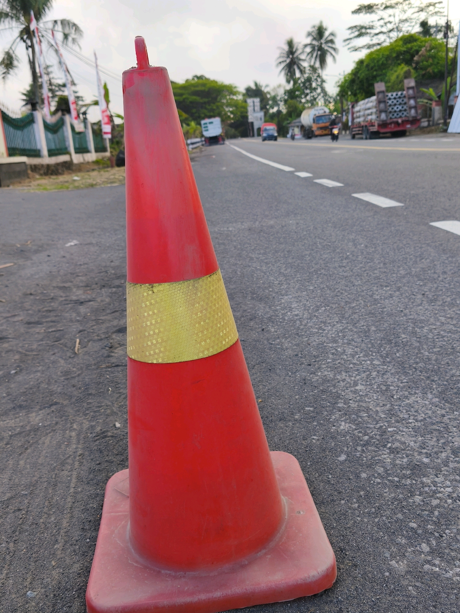 A red traffic cone with a yellow reflective band is positioned on the side of a two-lane asphalt road in Pringsurat, Indonesia. The cone is in the foreground, taking up a significant portion of the left side of the frame. The road has white dashed lane markings. In the background, a fence with green trim and several Indonesian flags can be seen on the left, likely indicating a residential or institutional area. Further down the road, several vehicles are visible, including a large truck with circular objects stacked on its flatbed, a cement truck, a smaller truck, and a motorcycle. The environment appears to be outdoors with a slightly overcast sky and lush green trees in the distance. The ground around the cone is also asphalt, with some debris.