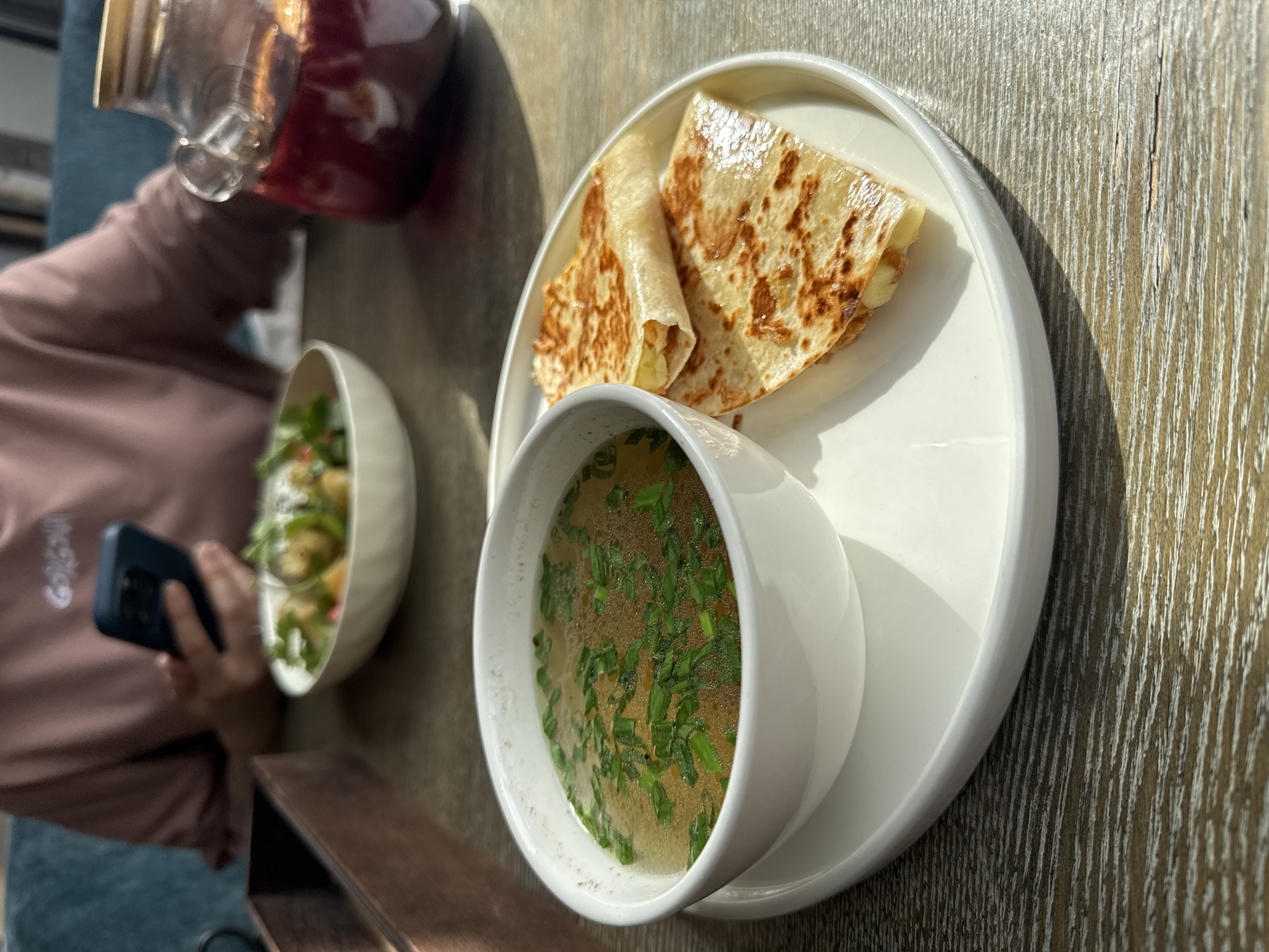 This media file captures a bright, sunlit meal scene set on a textured wooden table in Павловка, Russia. The central focus is a white round plate holding two main food items. On one side of the plate is a small, pristine white bowl filled with a clear broth soup, richly garnished with fresh green chives floating on the surface. Next to the soup, also on the white plate, are two pieces of a golden-brown, grilled flatbread, possibly a cheese quesadilla, folded into triangles, with melted cheese peeking out.
To the left of the main plate, a person is partially visible. She is wearing a light pink long-sleeved top and blue jeans. Her hand is holding a dark blue smartphone, suggesting she is interacting with it during the meal. Another white bowl, containing a fresh salad with various green leaves and possibly other ingredients, is partially visible closer to the person. Above the person's hand, a clear glass pitcher with a wooden lid, filled with a deep red beverage (possibly juice or kompot), is either being held or has recently been placed on the table.
The strong natural light indicates it is daytime, with distinct shadows cast on the table and the items, suggesting a sunny day, likely morning or early afternoon. The wooden table surface displays a prominent grain, contributing to a casual, possibly outdoor or semi-outdoor, dining ambiance. No discernible text is visible on any objects or clothing in the image.