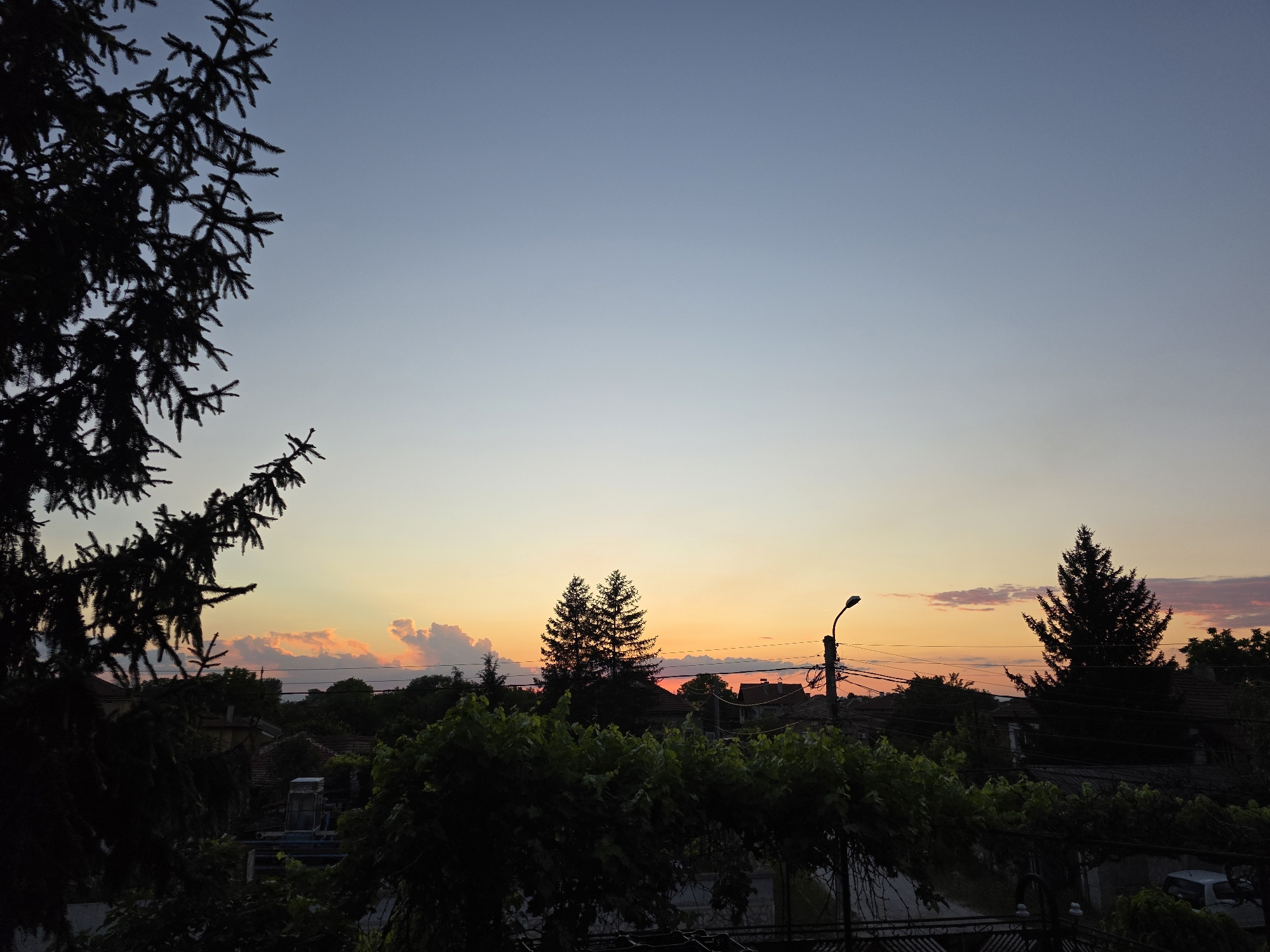 The image depicts a sunset view from an elevated position, likely a balcony or upper floor, in Yagodovo, Bulgaria. The sky transitions from a light blue at the top to orange and pink near the horizon. Scattered clouds are visible in the sunset colors. In the foreground, silhouetted trees and foliage fill the lower portion of the frame. Rooftops of houses are discernible amongst the trees, indicating a residential area. A utility pole with wires is visible against the sunset. A conifer tree stands to the right side of the frame. The lighting suggests the photo was taken during the evening, shortly after sunset.
