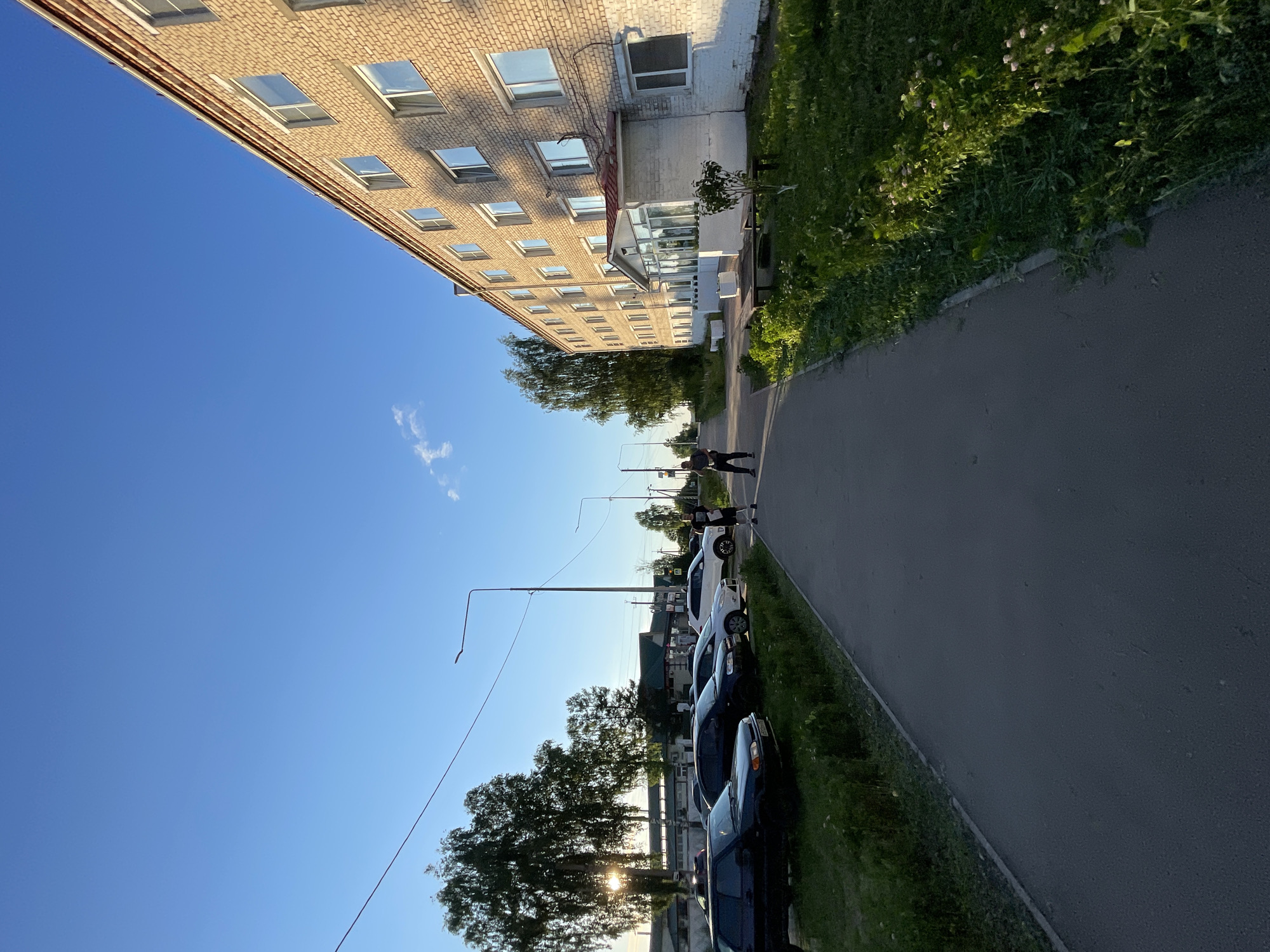The image depicts a residential building constructed with light-colored bricks, featuring multiple floors and numerous windows. A paved walkway runs alongside the building, bordered by a strip of green grass with scattered vegetation. To the left of the walkway, a row of parked cars is visible. Two individuals are standing on the walkway, facing away from the camera. The scene is set under a clear blue sky with a few wispy clouds, suggesting it is daytime. The overall environment appears to be an outdoor, urban setting. Based on the context provided, this scene is located in the city of Krasnye Baki, Russia.