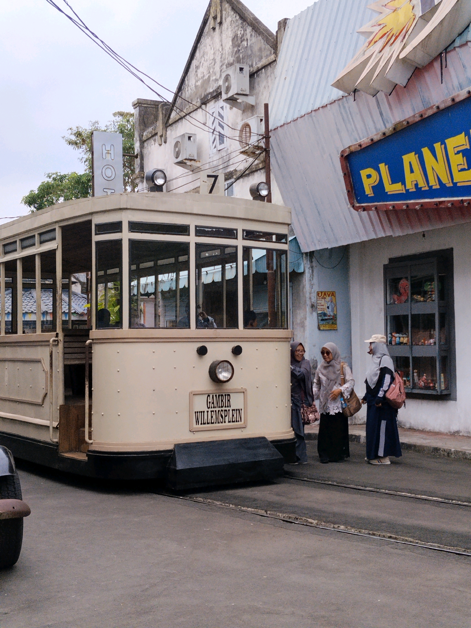 The image depicts a street scene in the city of Sumberrahayu, Indonesia, featuring a vintage tram and a group of people. The tram, light beige in color with black trim and large windows, is positioned on tram tracks in the foreground. A sign on the side of the tram reads "GAMBIR WILLEMSPLEIN". To the right of the tram, three women are standing on the sidewalk. The woman on the far left wears a dark hijab and a patterned bag. The middle woman has a light gray hijab, sunglasses, and a brown bag. The woman on the right wears a light hijab, a cap, and a pink backpack. Behind the women, a building with a white facade displays a blue sign with yellow lettering that reads "PLANET". A decorative burst of yellow and red is above the "PLANET" sign. Further back, another building, partially visible, has a sign that reads "HOT". The overall environment appears to be an urban setting with a slightly overcast sky.